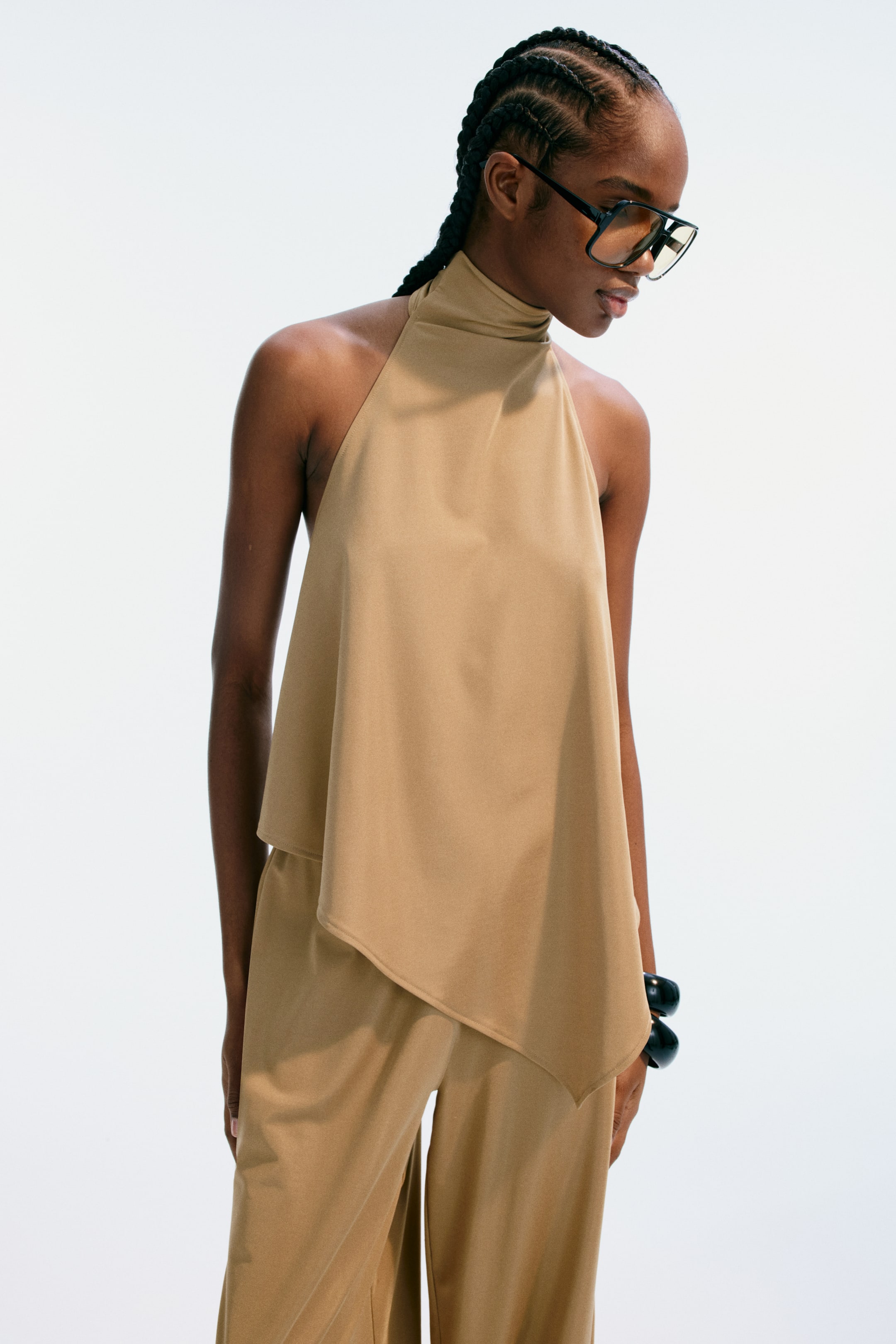 Visualizza immagine più grande: Un completo a due pezzi color cammello presenta un top halter a collo alto con un orlo asimmetrico e fluente, sovrapposto a pantaloni a gamba larga coordinati. Il completo è leggermente inclinato a destra, mettendo in risalto il suo tessuto drappeggiato e la vestibilità morbida.