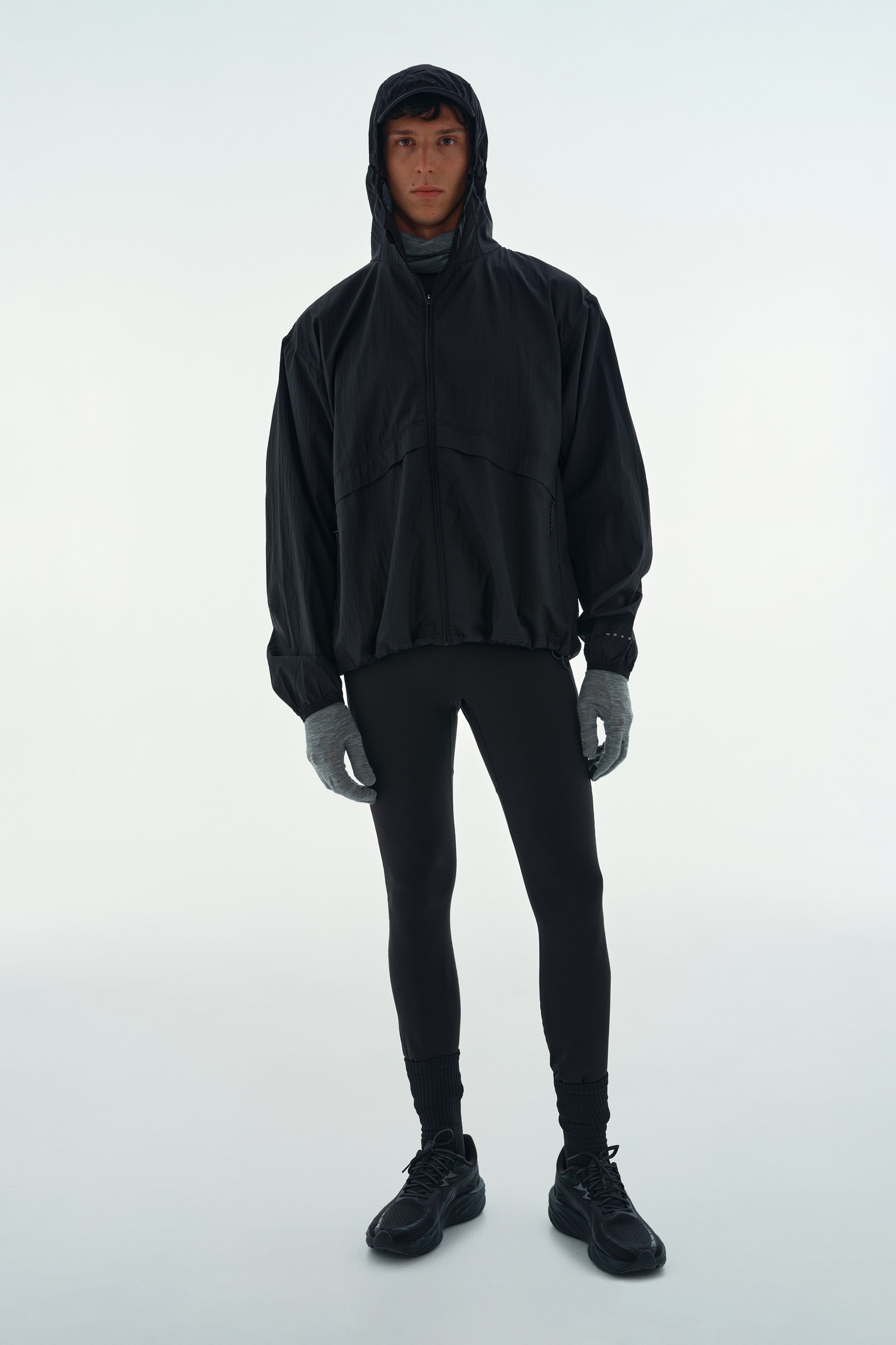 View larger image: A black, lightweight running jacket with a relaxed fit is being worn by the model, featuring a raised hood, dropped shoulders, and elasticated cuffs. The jacket is styled with black athletic tights, grey gloves, and black running shoes.