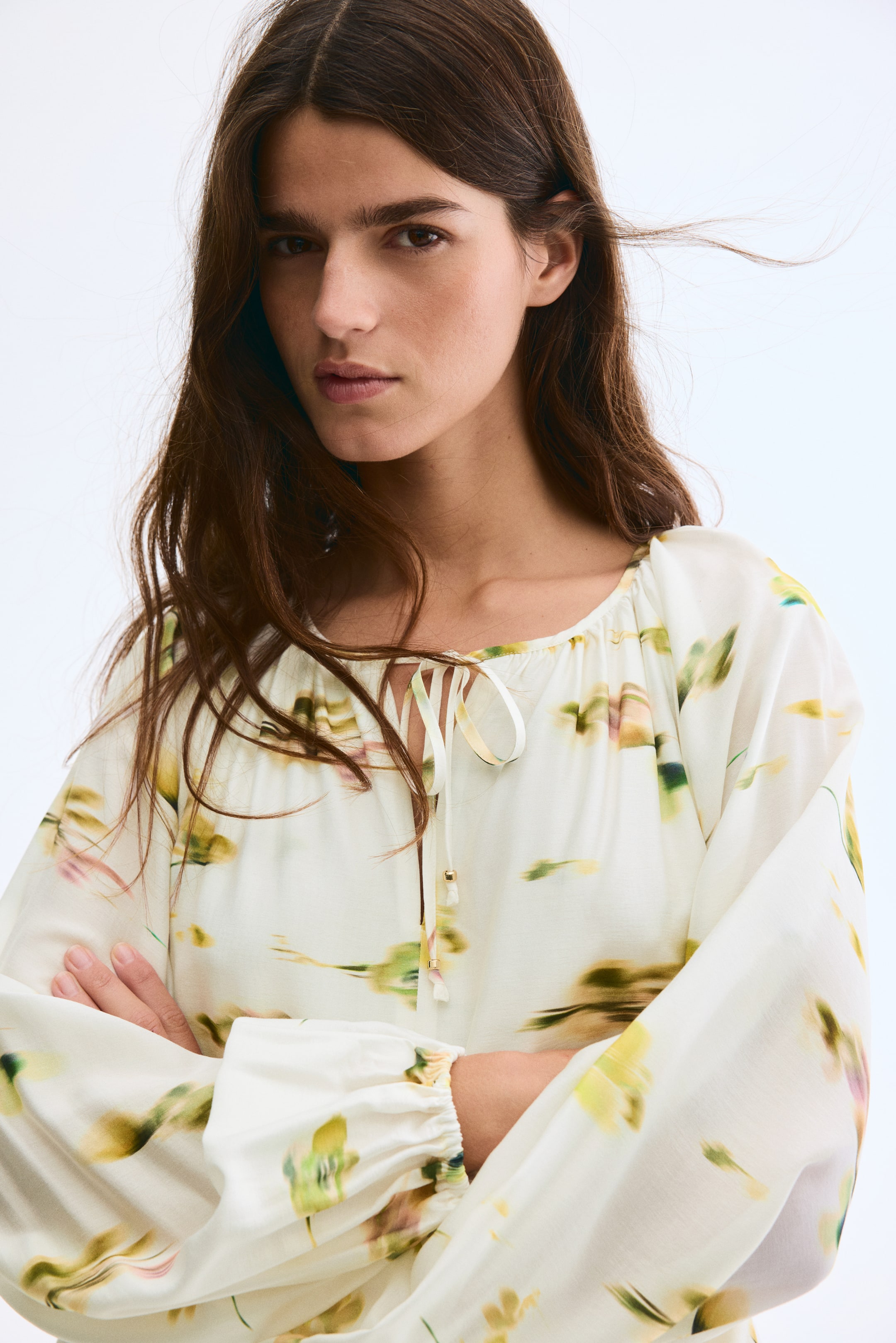 Ampliar la imagen: Una blusa de color crema presenta un patrón floral abstracto en tonos de verde, amarillo y un sutil rosa. Está diseñada con un escote redondeado recogido, un cierre frontal con lazo y extremos pequeños con cuentas, y mangas largas y voluminosas con puños recogidos.