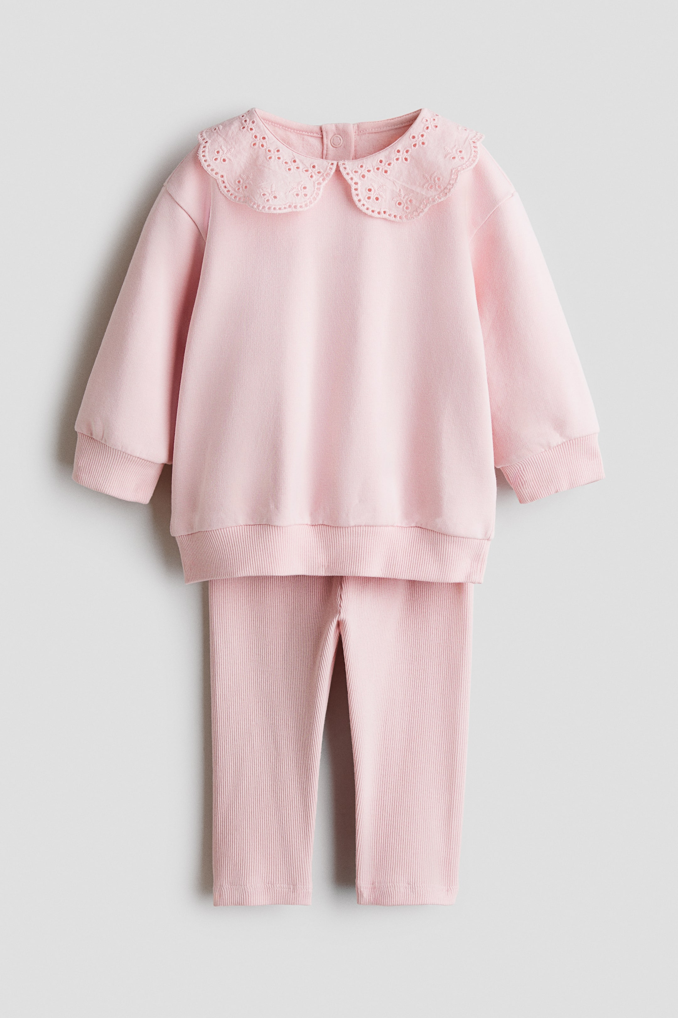 Visa större bild: Ett ljusrosa tvådelat babyset visas framifrån och består av en långärmad sweatshirt med en stor krage med vågad kant, dekorerad med broderie anglaise-broderi. Sweatshirten, med nedhasade axlar samt ribbade muddar och nederkant, matchas med ljusrosa finribbade leggings i samma färg.