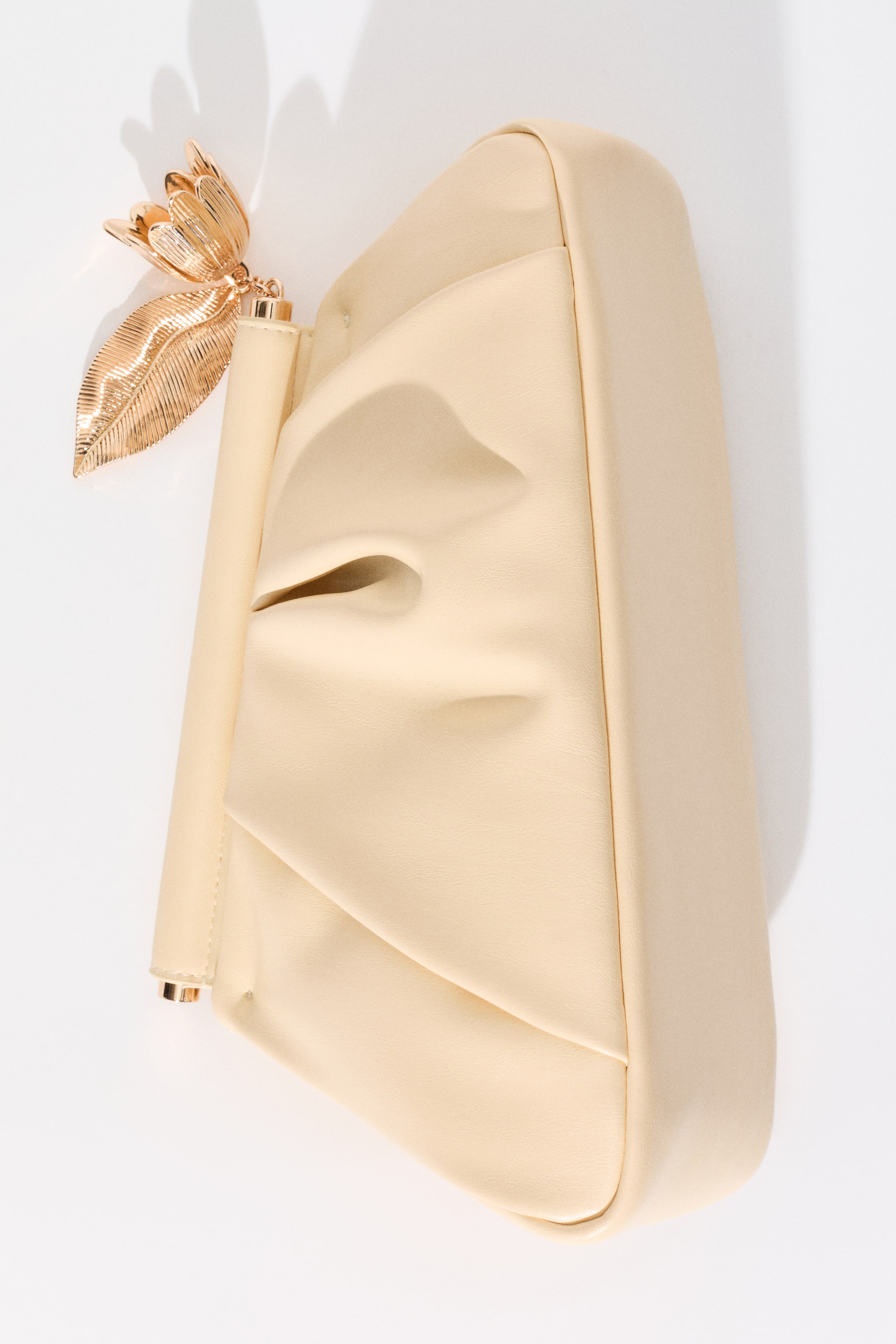 View larger image: A cream-coloured clutch bag is resting on its side, angled upwards, showcasing a gathered and pleated soft material front. A rigid cylindrical bar forms its top closure, adorned with a golden metal charm featuring a detailed leaf and a multi-petalled flower hanging from it.