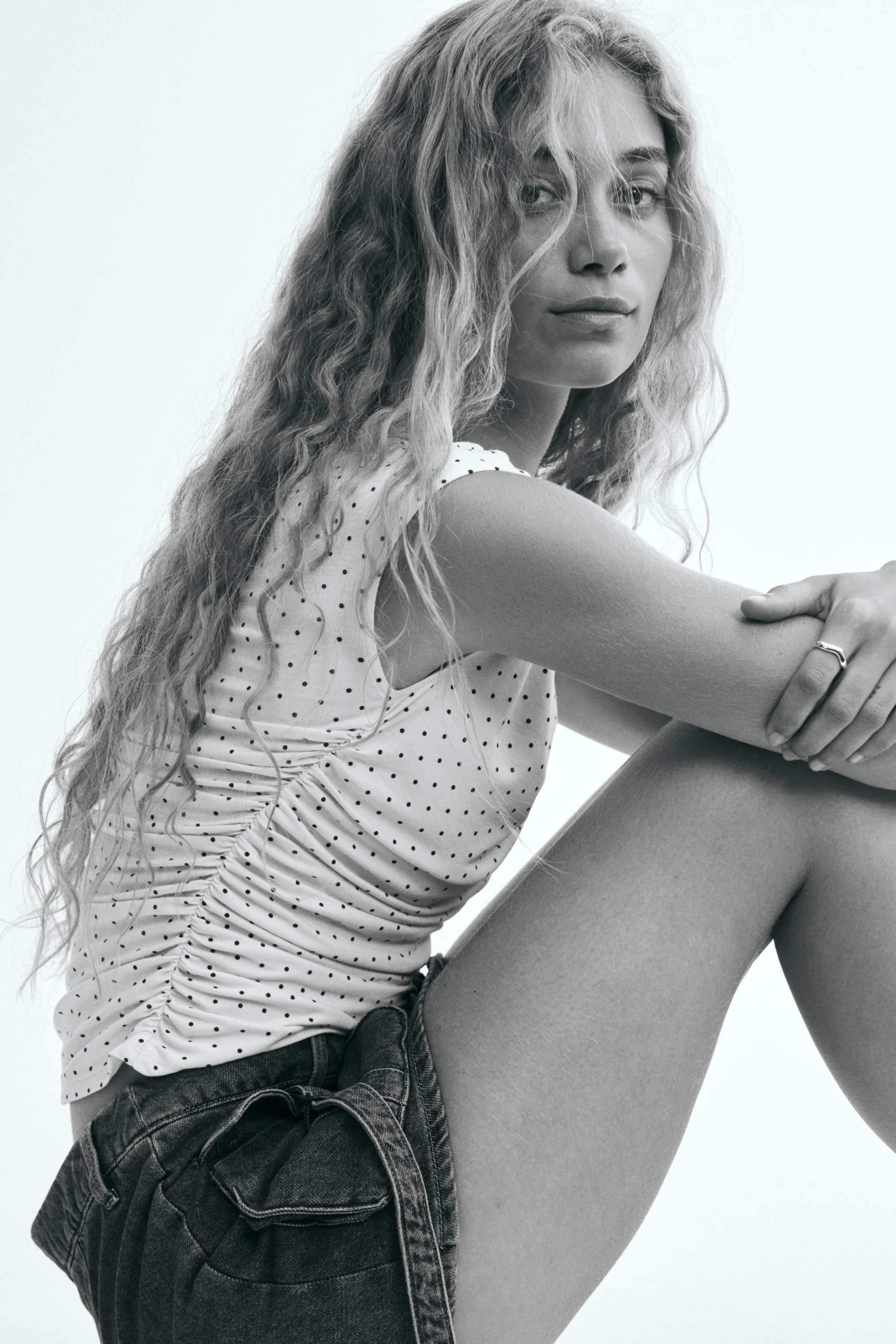 View larger image: A fitted, polka-dot patterned top featuring a gathered side seam and cap sleeves is being worn by the model, who is sitting in profile. The top is styled with dark denim shorts.