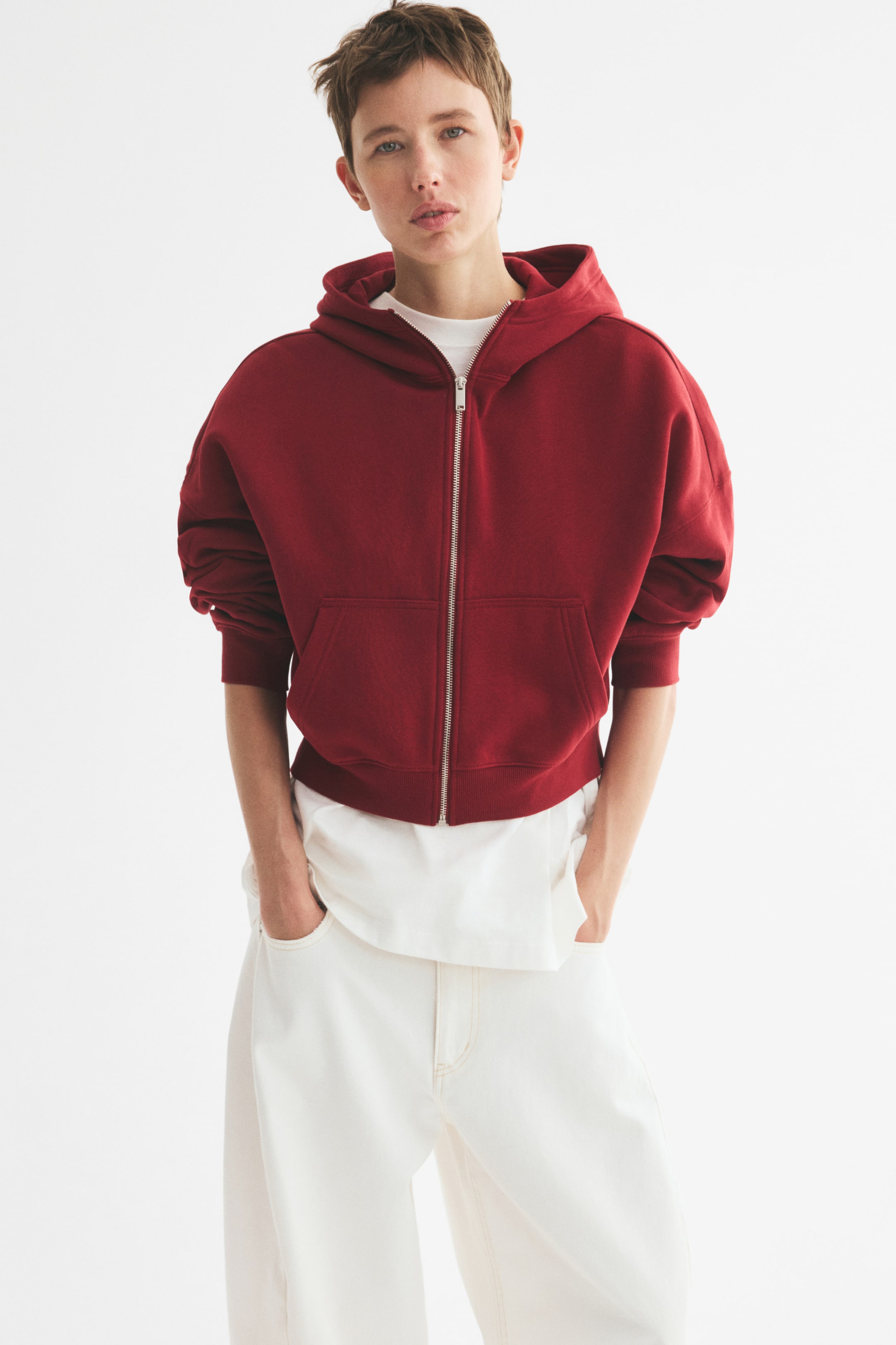 View larger image: The model is wearing a loose-fit, dark red hoodie with a fully zipped front, a lined hood, dropped shoulders, patch front pockets, and ribbing at the cuffs and hem. They have layered it over a white t-shirt and are resting their hands in the front pockets.
