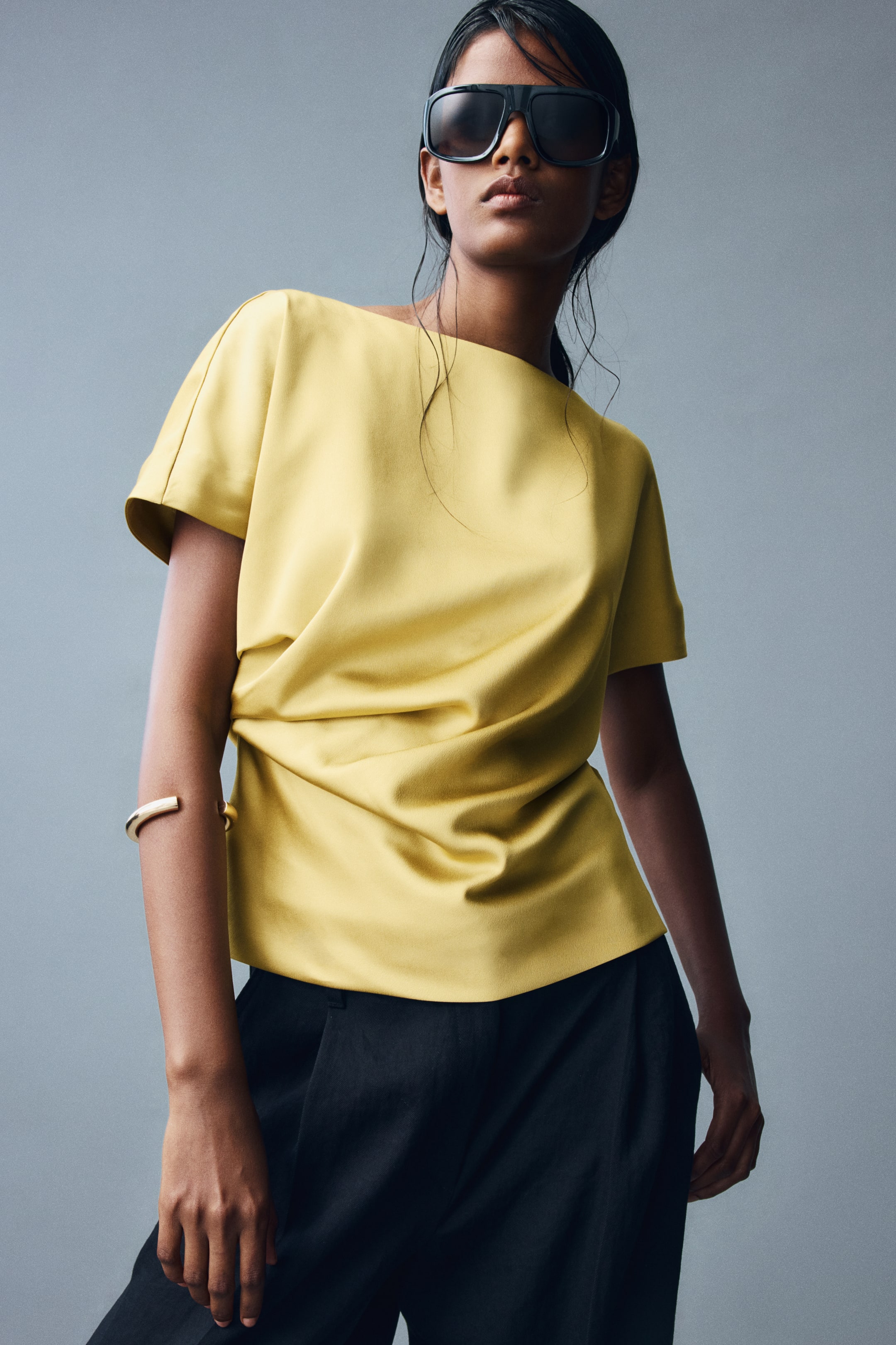 View larger image: A yellow satin blouse featuring a boat neck, cap sleeves, and draped pleating at one side of the waist is being worn by the model. The blouse is styled with black trousers, sunglasses, and a gold cuff on the upper arm.