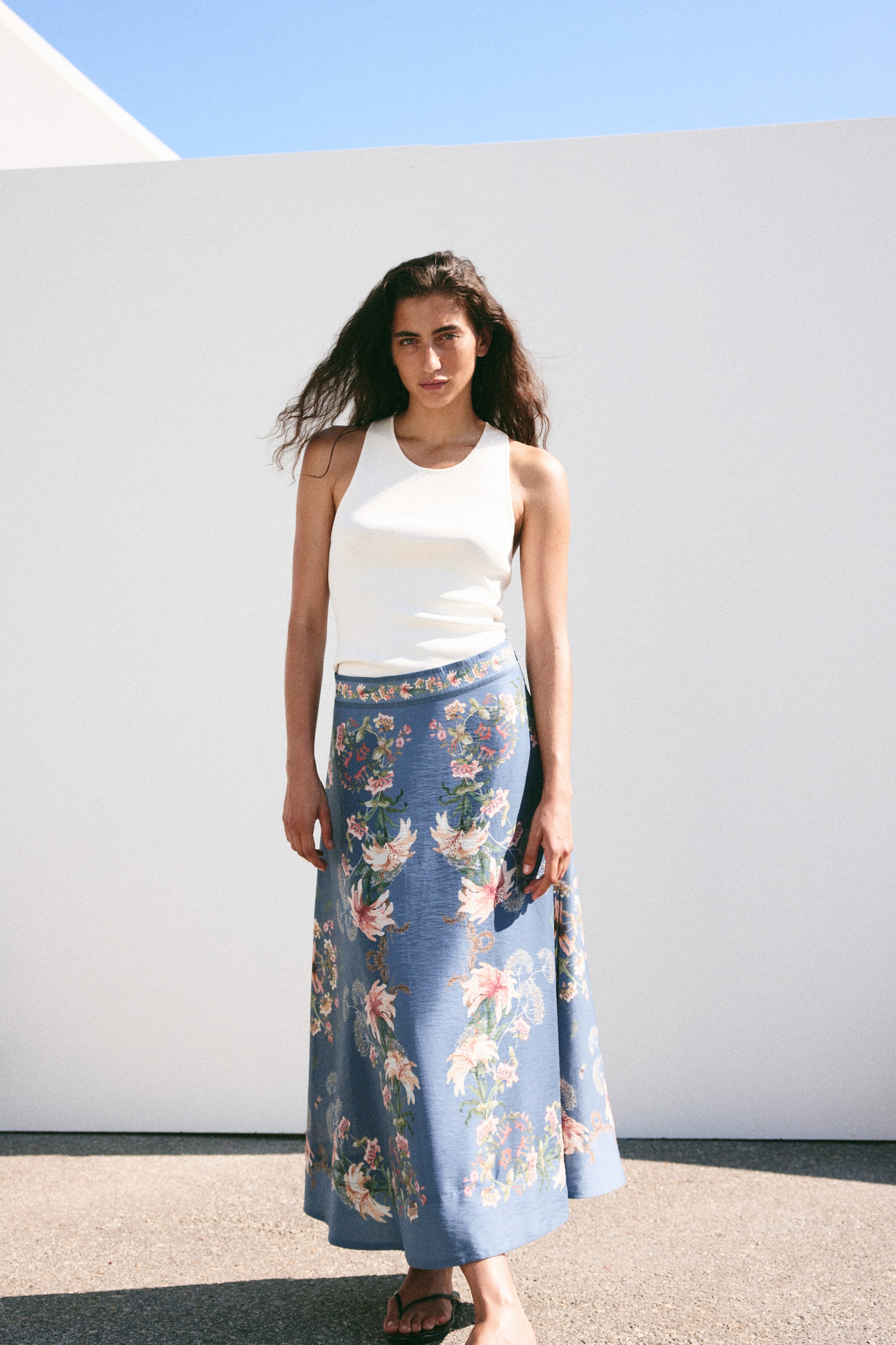 Grotere afbeelding bekijken: Een lange blauwe rok met een veelkleurig bloemenpatroon en een uitlopende snit wordt door het model gedragen. Ze wordt gecombineerd met een witte tanktop.