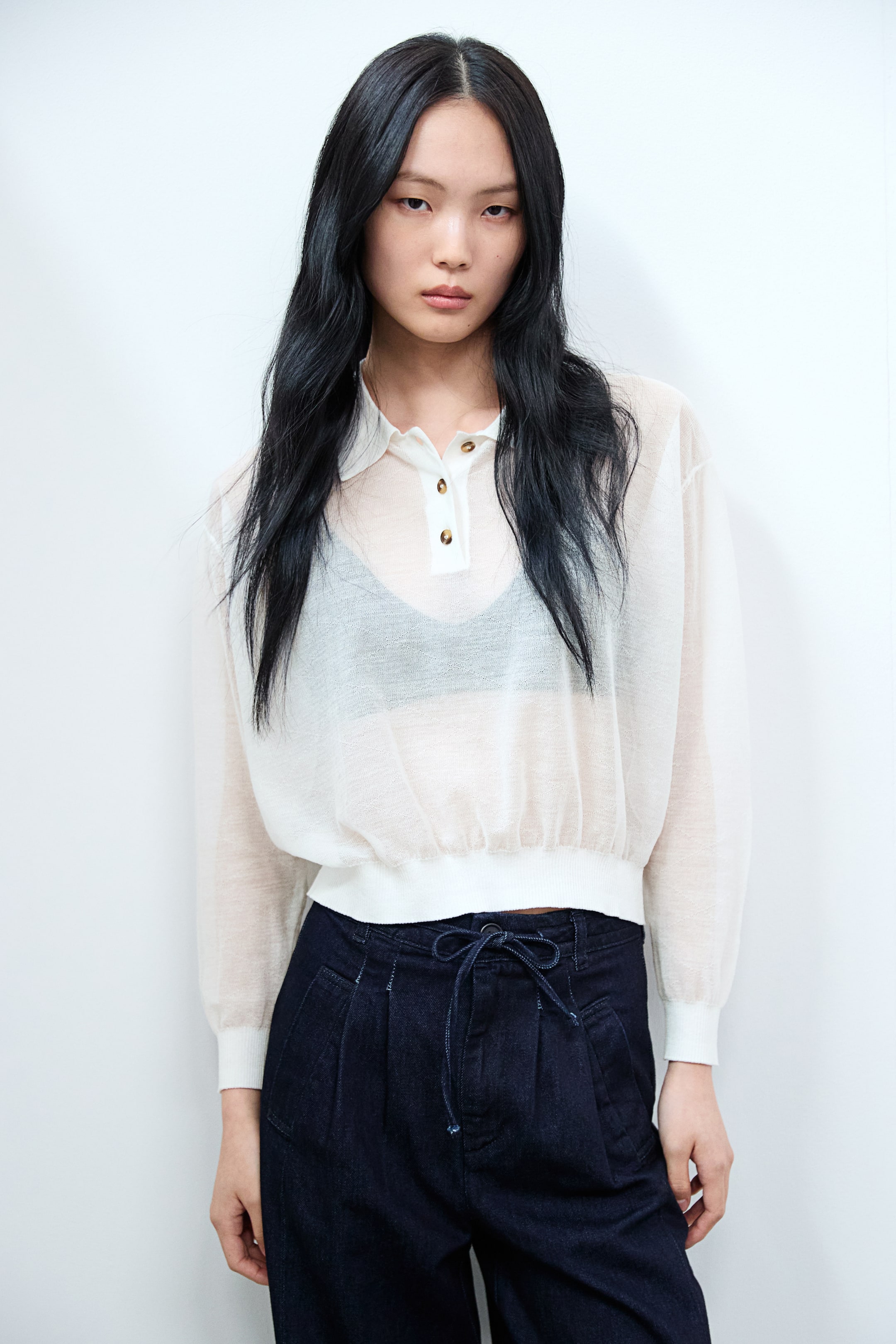 拡大画像を表示: シアーなオフホワイトのポロトップは、ブラウンのボタンが付いた2ボタンのプラケット、リブ付きカフスの長いボリューム袖、そしてエラスティックリブの裾が特徴で、ブラウゾンシルエットを作り出しています。ダークなブラジャーが下に着用されており、トップの透け感のある生地を際立たせています。