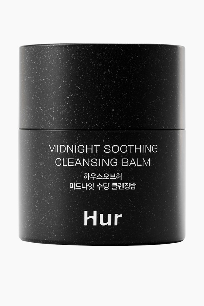 En sort cylindrisk rensebalsam krukke præsenterer sin frontetiket. Krukken har et plettet mønster af små hvide pletter over dens overflade, med hvid tekst, der viser "MIDNIGHT SOOTHING CLEANSING BALM," koreansk skrift og mærkenavnet "Hur." - 1