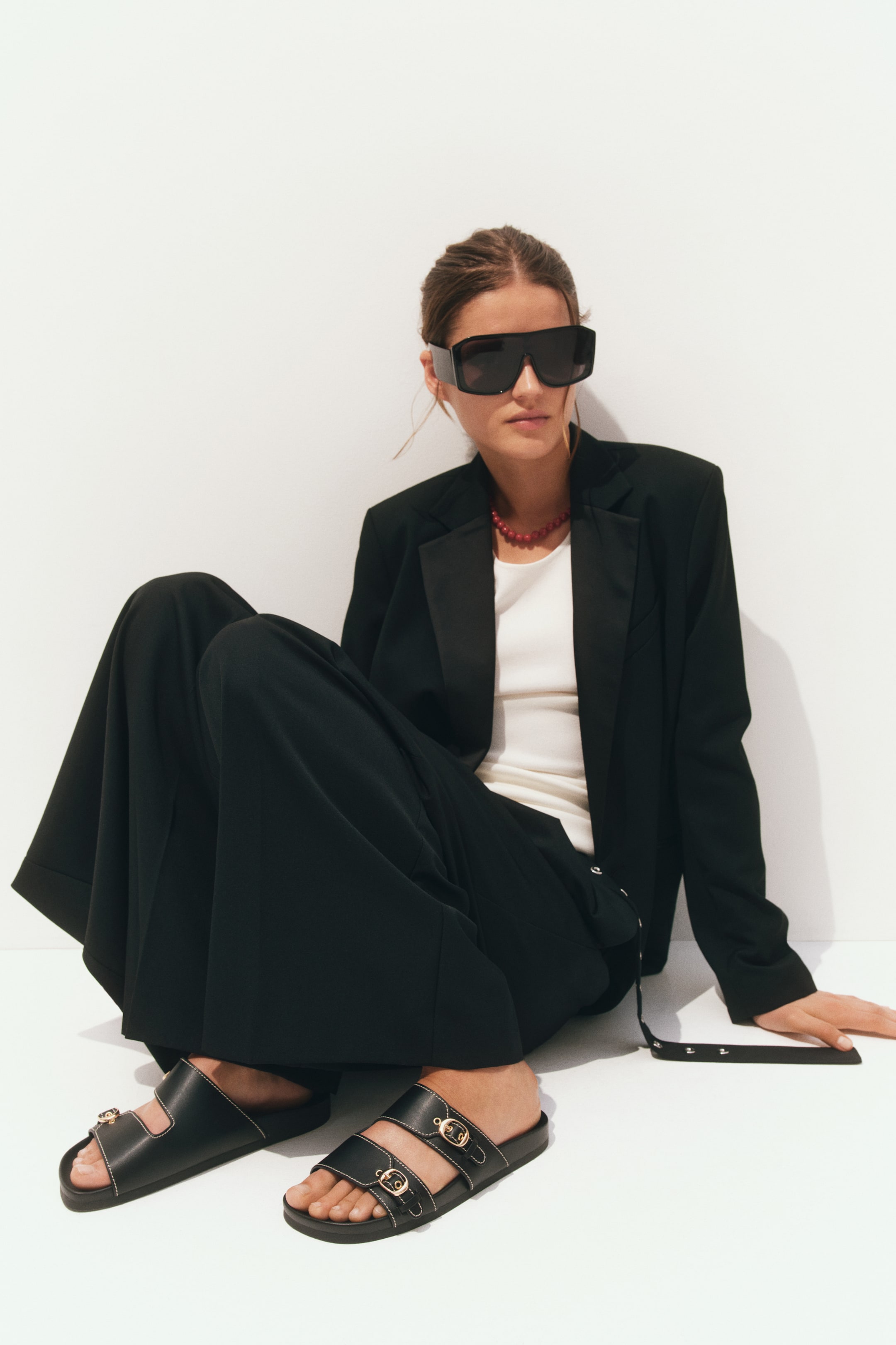 Ver imagen más grande: Sandalias de tela revestida, con punteras oblicuas y dos correas ajustables para el pie con hebillas de metal en tono dorado, son llevadas por la modelo. Las correas negras están detalladas con costuras blancas.