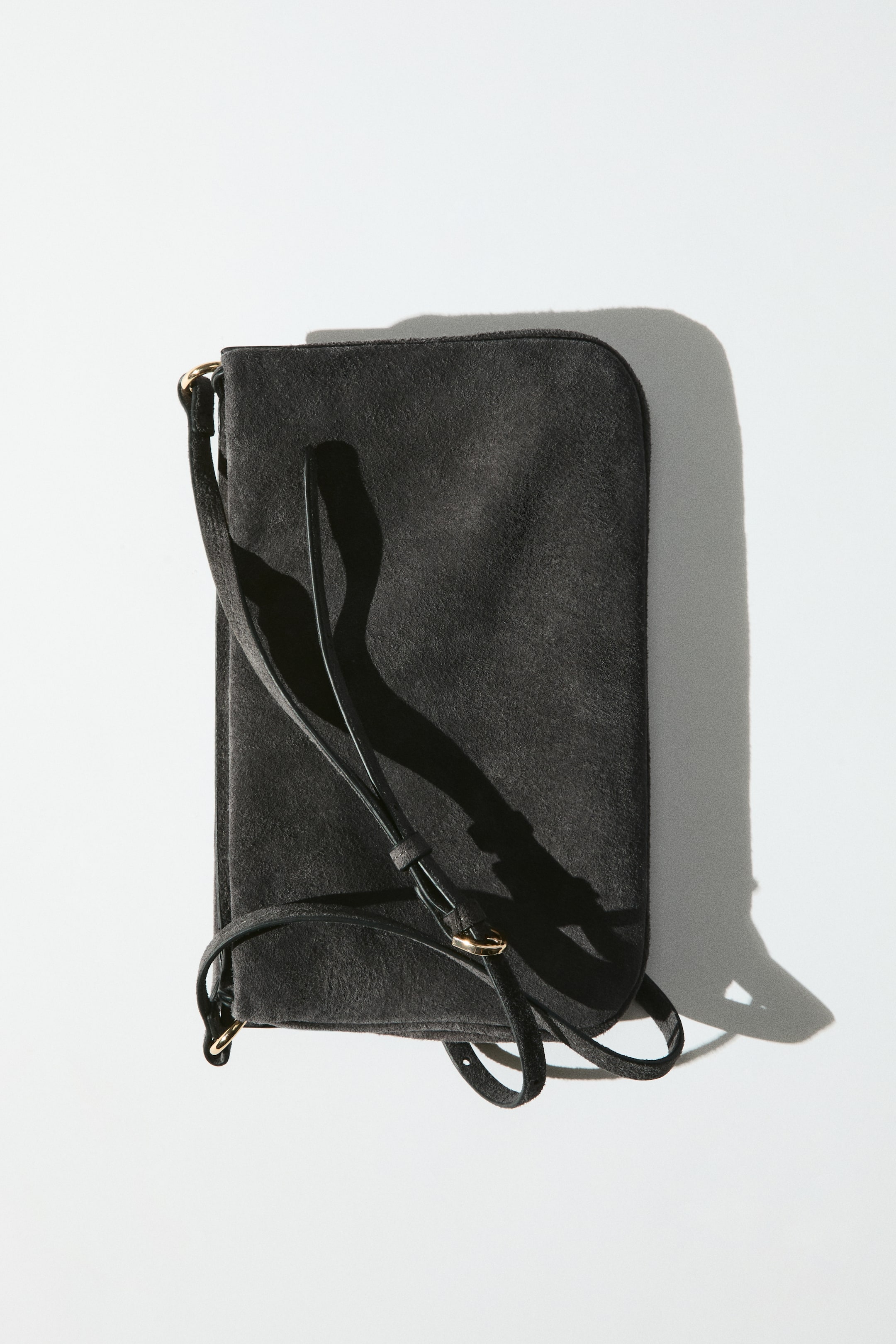 Zobraziť väčší obrázok: Čierna semišová crossbody kabelka leží naplocho a zobrazuje svoju prednú stranu s mäkkou, štruktúrovanou povrchovou úpravou a obdĺžnikovým tvarom. Úzky, nastaviteľný ramenný popruh so zlatistou prackou sa preťahuje cez prednú stranu kabelky.