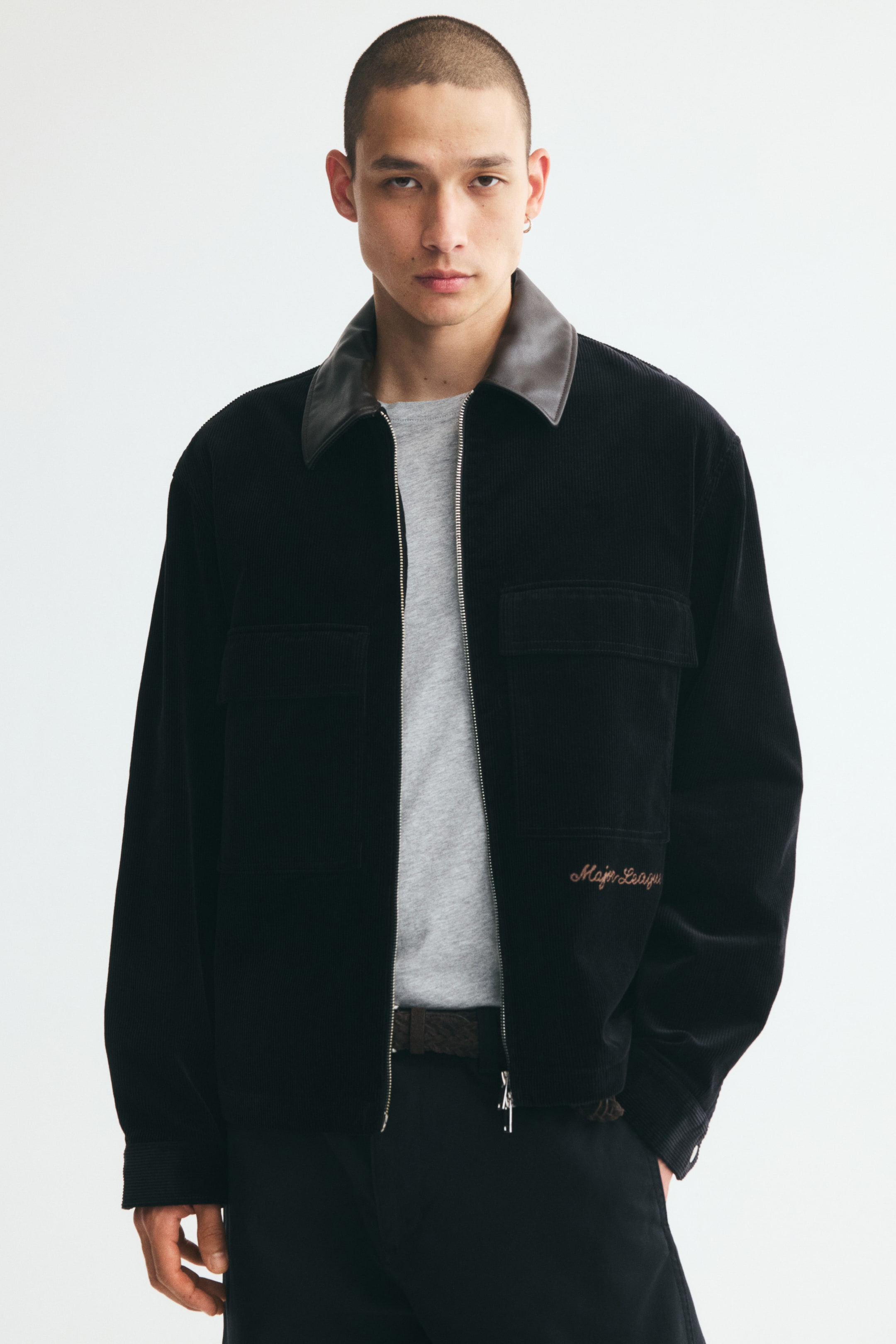 Grösseres Bild ansehen: Eine schwarze Cordjacke mit kontrastierendem braunem Lederkragen wird vom Model über einem grauen T-Shirt und dunklen Hosen getragen. Die Jacke verfügt über einen silberfarbenen Frontreissverschluss, grosse aufgesetzte Brusttaschen mit Patten und eine aufgestickte Schriftzug-Details auf der linken unteren Seite.
