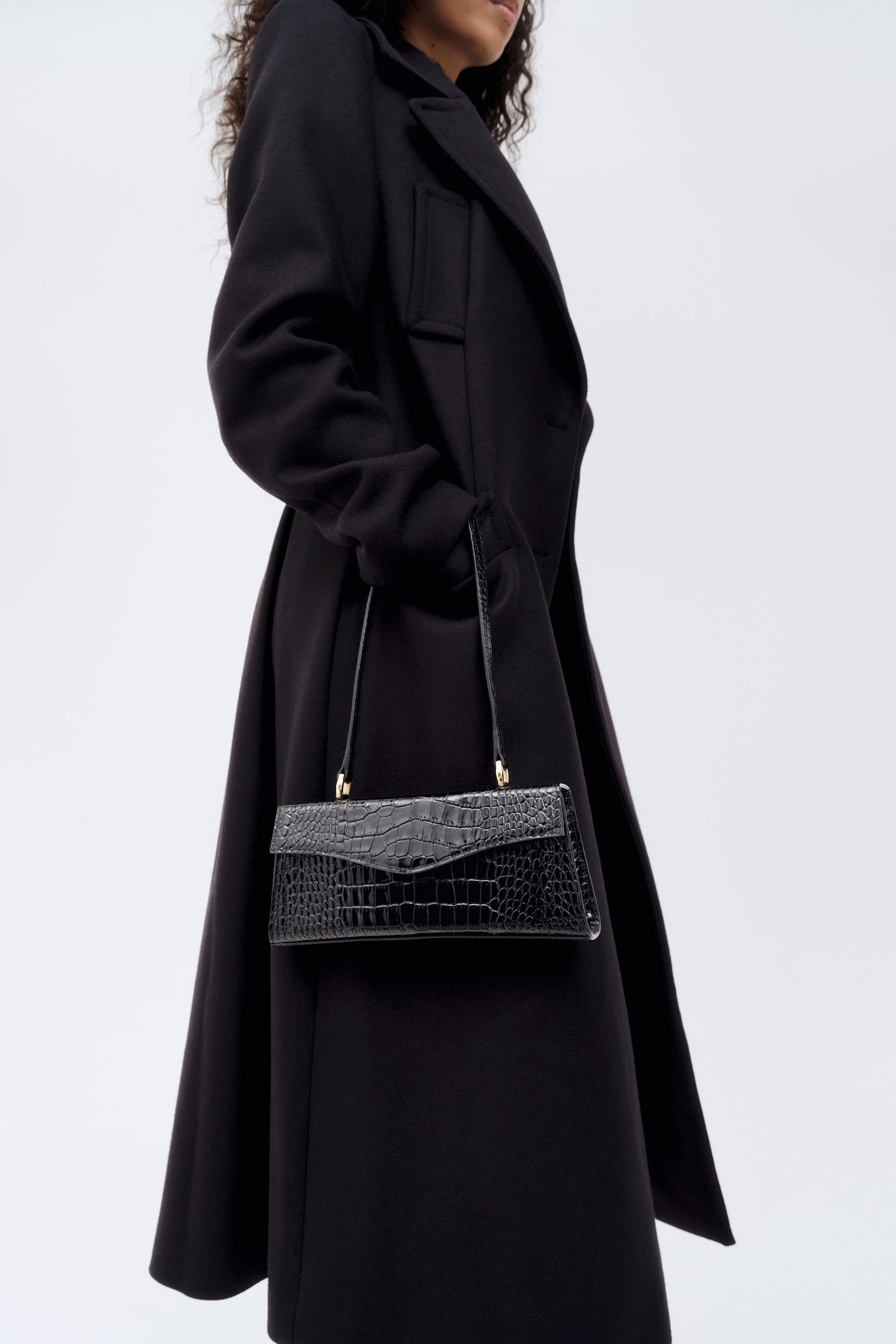 View larger image: A black calf-length coat in a wool-blend weave is shown from a side profile facing right, featuring a large collar, wide peak lapels, and a structured silhouette. The model is holding a black croc-embossed shoulder bag with gold-tone hardware while keeping one hand tucked into the coat's pocket.