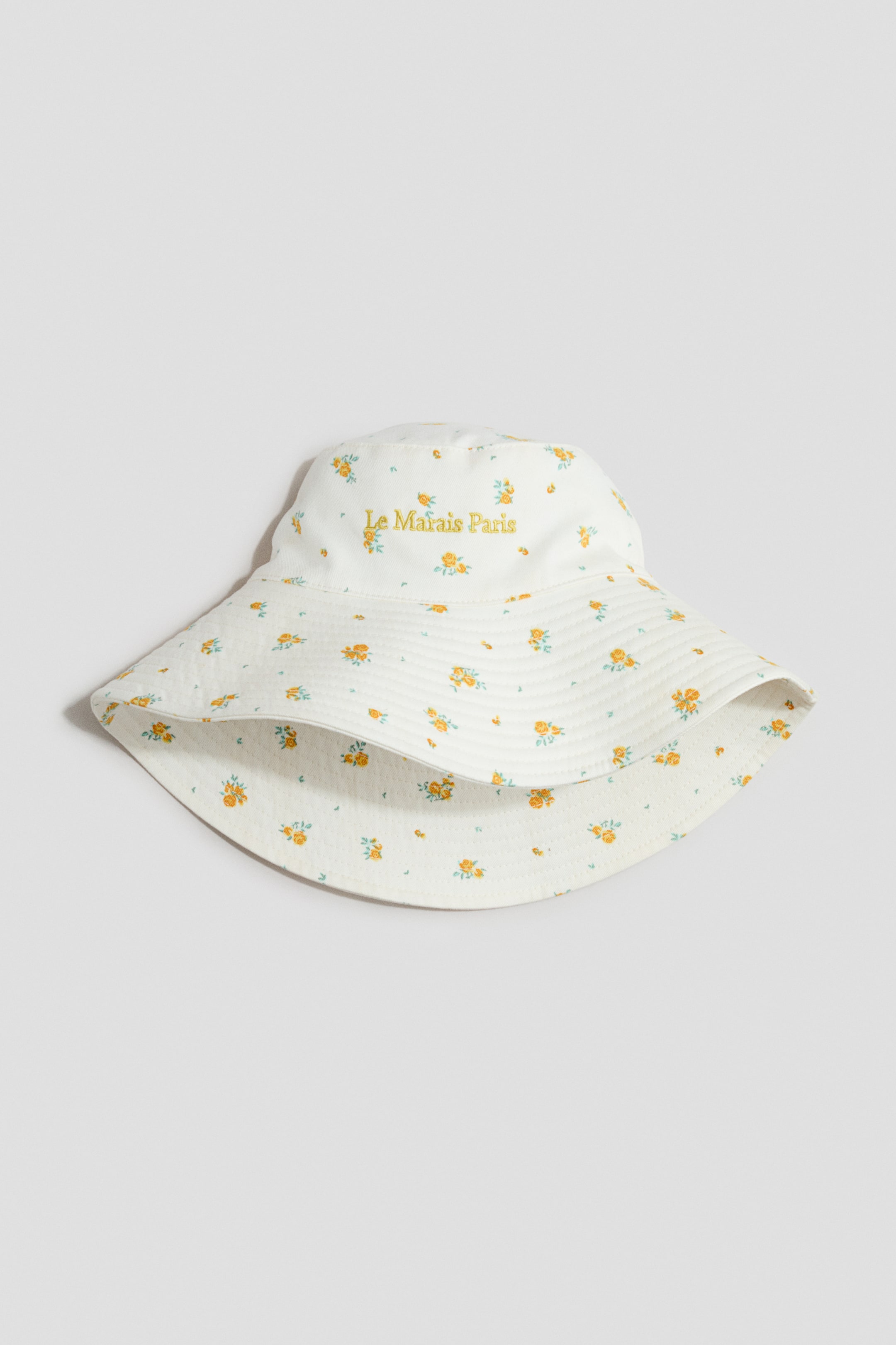 View larger image: A white cotton twill sun hat is displayed from the front, showcasing a yellow and green floral design across its outer surface and visible inner lining. It is crafted with a broad, horizontally stitched, sloping brim, with "Le Marais Paris" embroidered in yellow on the crown.