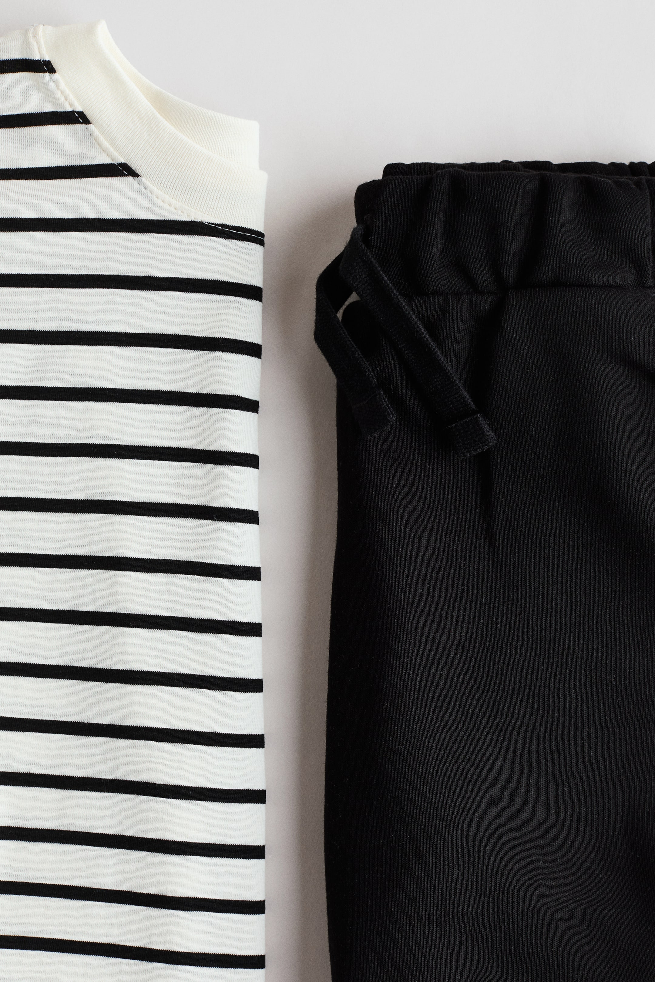 Größeres Bild anzeigen: Ein T-Shirt und Shorts-Set wird präsentiert, das ein weißes T-Shirt mit schwarzen horizontalen Streifen und einem runden, gerippten Halsausschnitt zeigt, neben schwarzen Shorts mit einem elastischen Kordelzugbund.