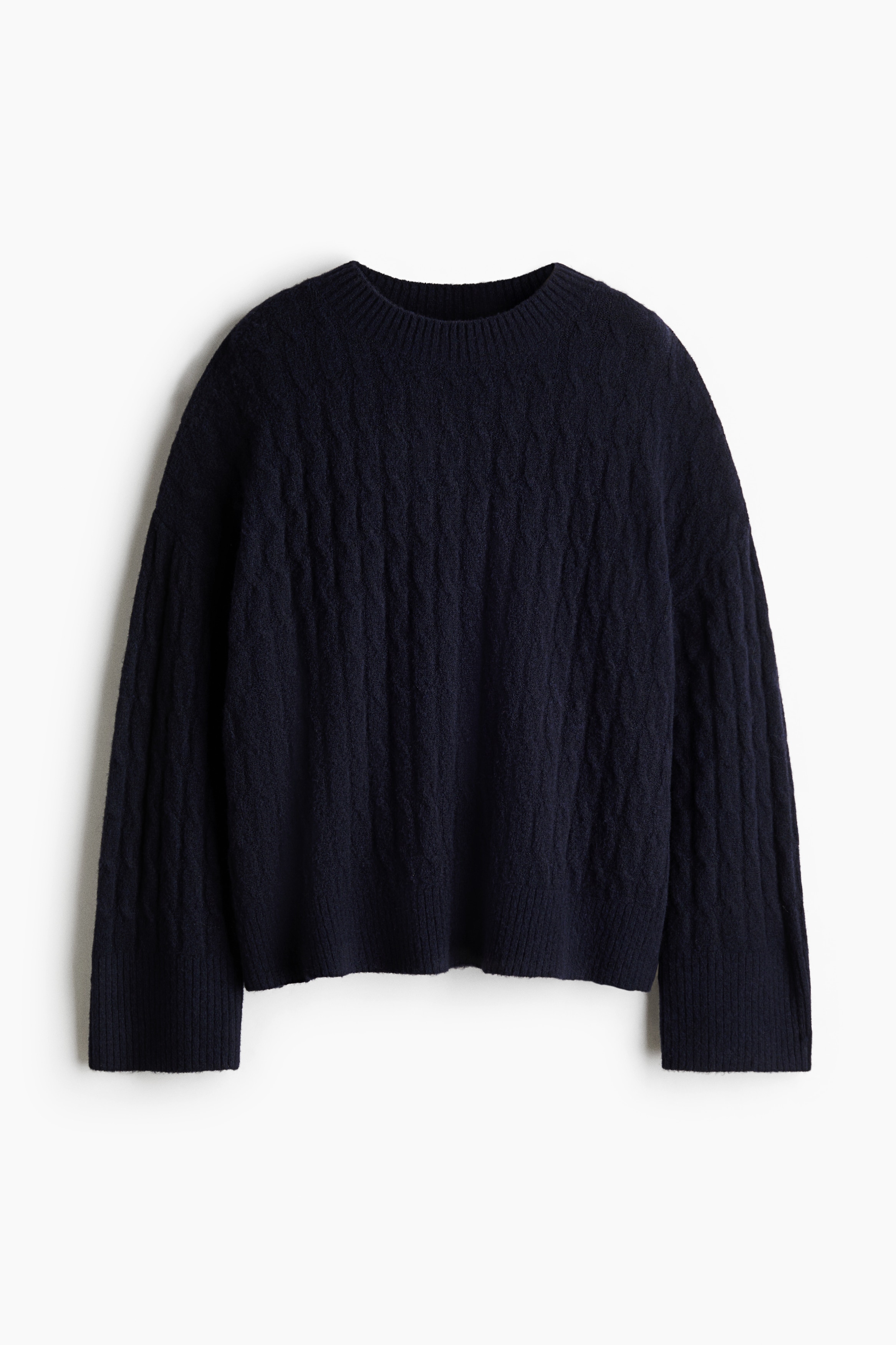 A dark navy, loose-fit cable-knit jumper featuring dropped shoulders and long sleeves with ribbed cuffs is being worn by the model. The jumper is paired with light-coloured shorts as the model sits in profile.