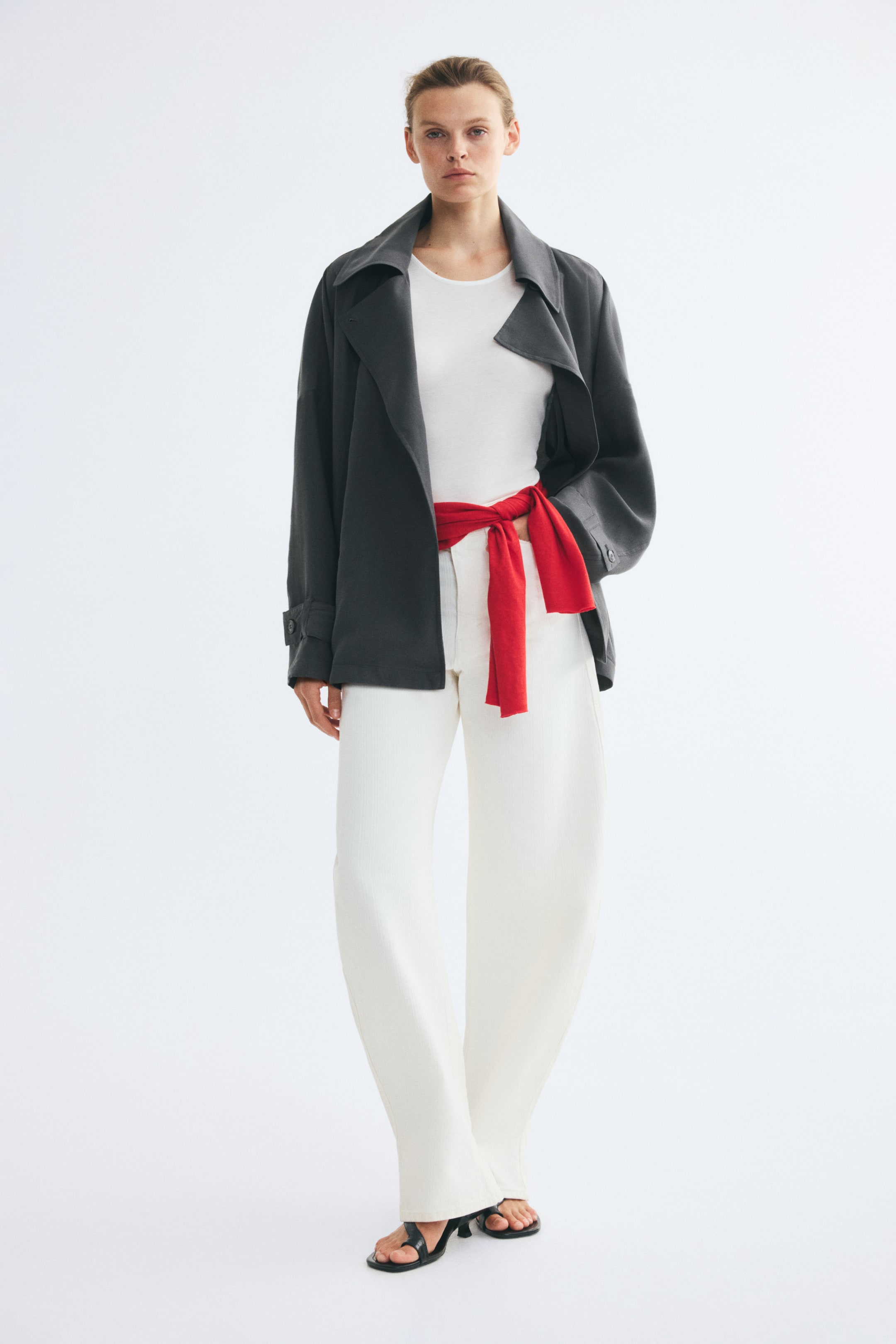 View larger image: A short, dark grey oversized trench coat is worn open by the model, featuring a large collar, wide notch lapels, and button tabs at the cuffs. The coat is layered over a white top and matching white trousers, accented by a bright red knit sweater tied around the waist.