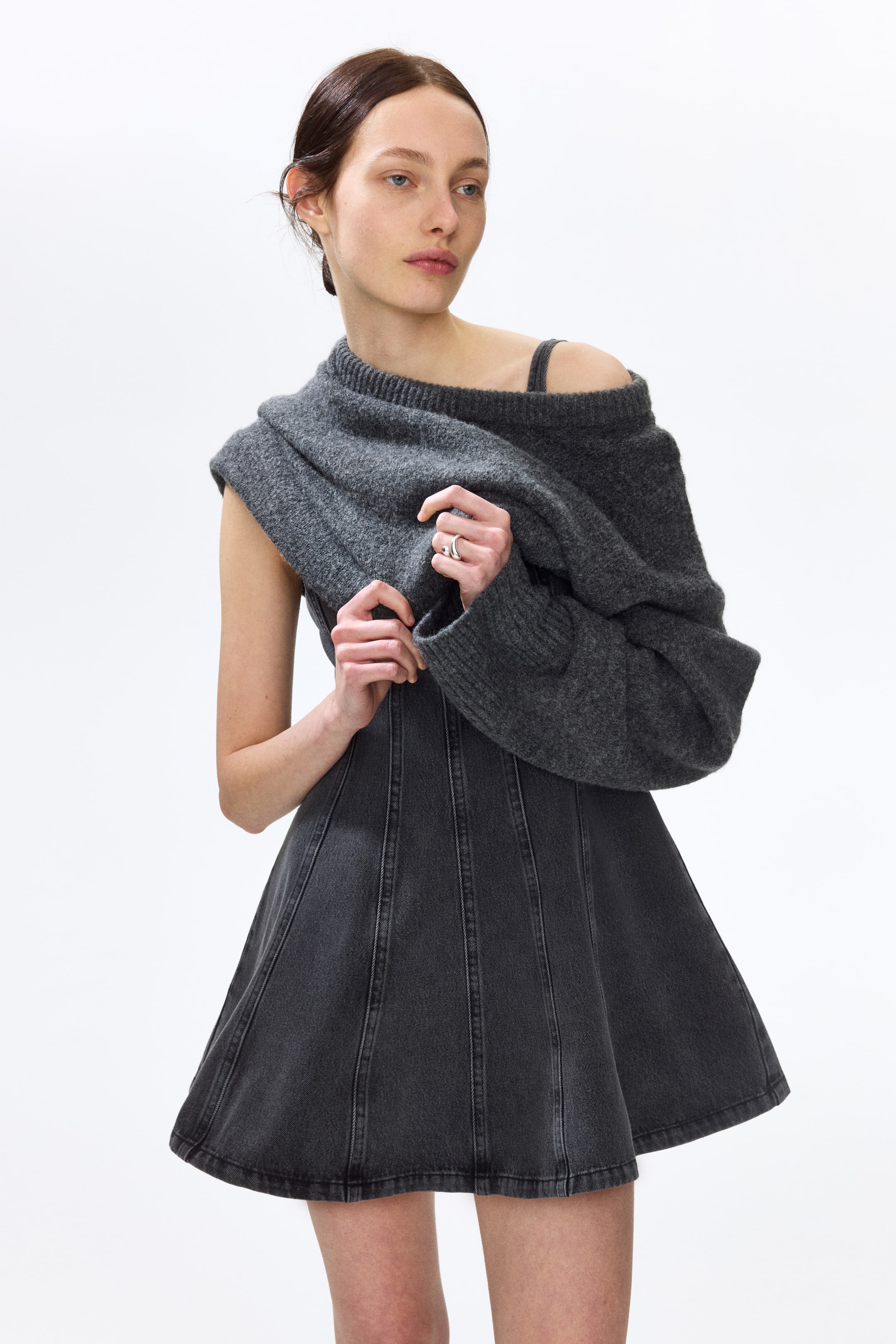 拡大画像を表示: ダークグレーのニットセーターが片方の肩と腕にかかり、リブ付きのクルーネックとリブ付きのカフスが特徴です。ダークグレーのデニムAラインミニスカートの上に重ねられ、縦のシームディテールが施されています。