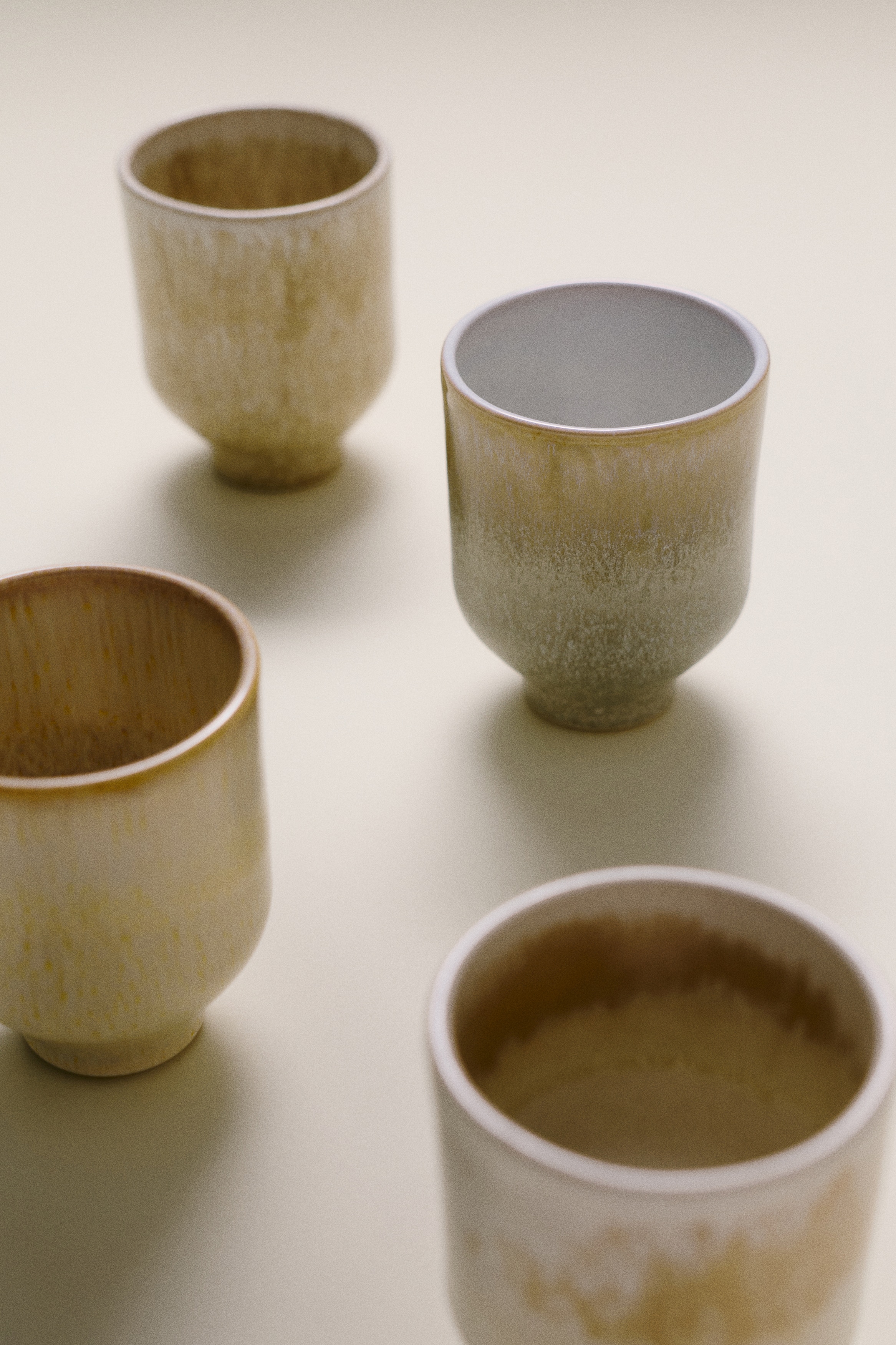 Patru cÄni ceramice fÄrÄ mÃ¢ner sunt aÈezate Ã®ntr-un rÃ¢nd orizontal, orientate spre faÈÄ pentru a evidenÈia designurile lor unice. Fiecare canÄ are o bazÄ tip piedestal conicÄ Èi o glazurÄ distinctÄ, organicÄ, Ã®n nuanÈe pÄmÃ¢ntii de crem, bej Èi gri deschis, cu finisaje Ã®n degrade texturat sau cu efect de scurgere.