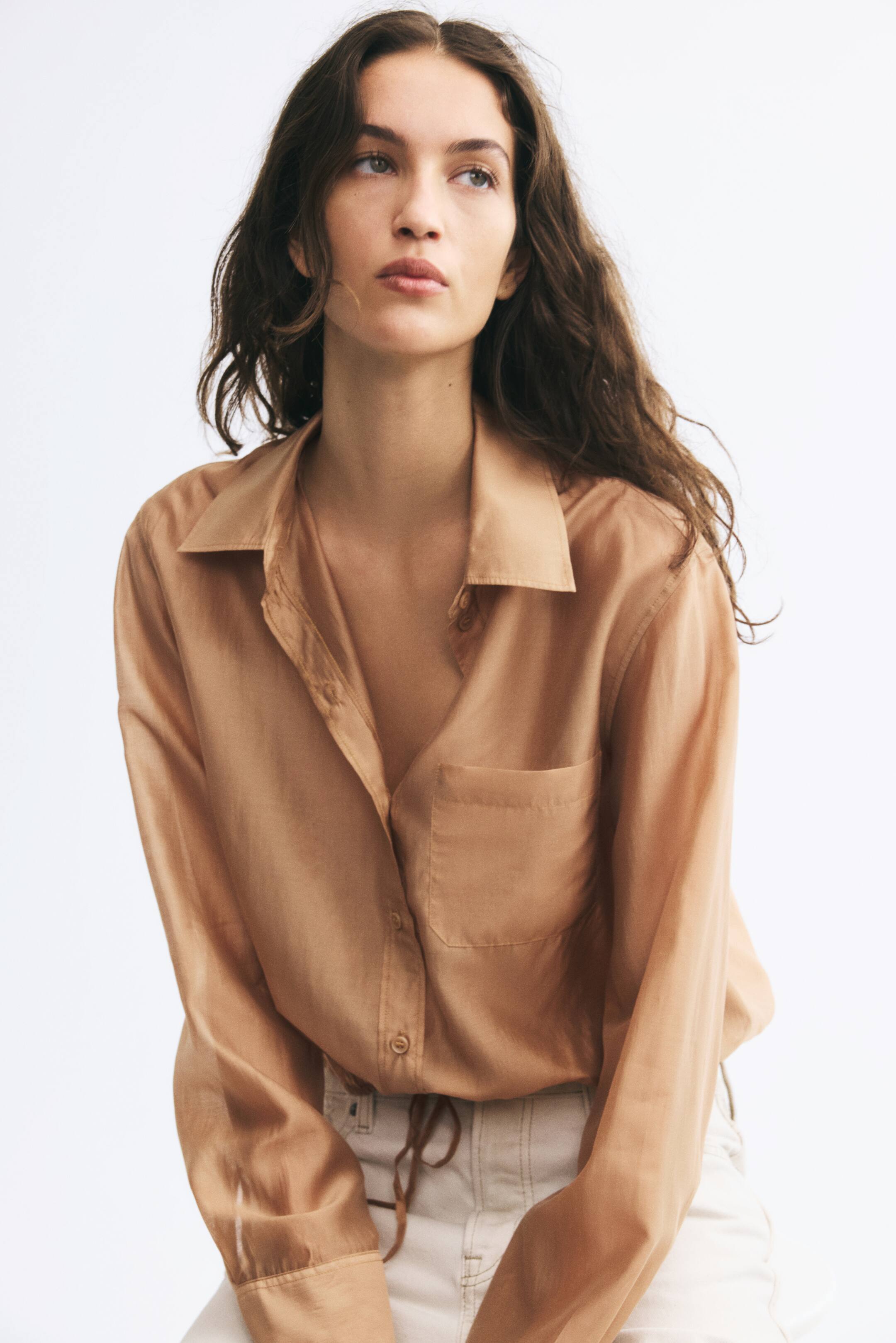 View larger image: A bronze-colored, long-sleeved shirt with a subtle sheen is worn by the model. The shirt features a collar, a partially unbuttoned front, a single patch chest pocket, and a drawstring hem with ties draping over off-white trousers.
