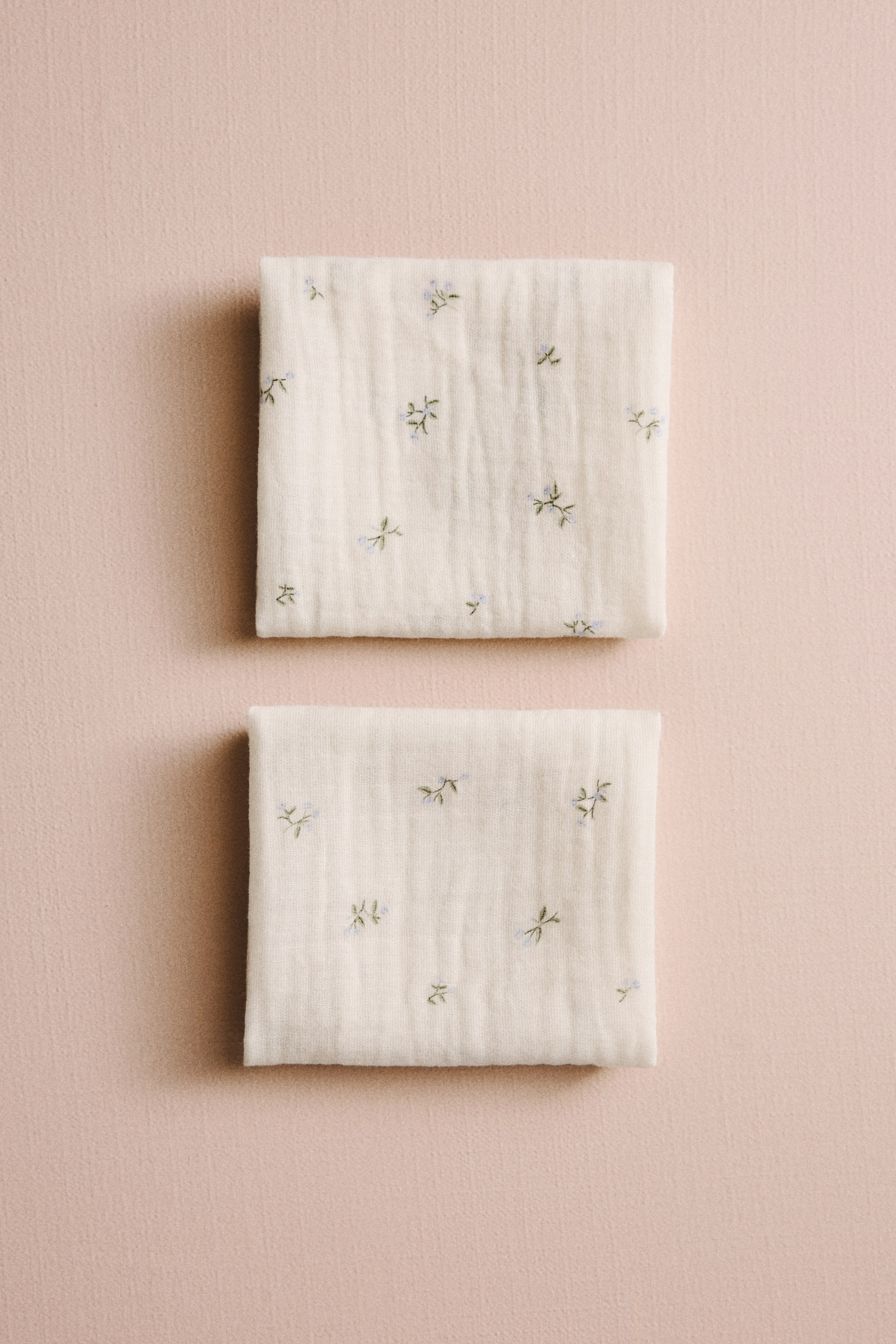 View larger image: Two cream-colored muslin cloths are folded into rectangles. They feature a textured, crinkled surface with scattered blue and green floral embroidery.