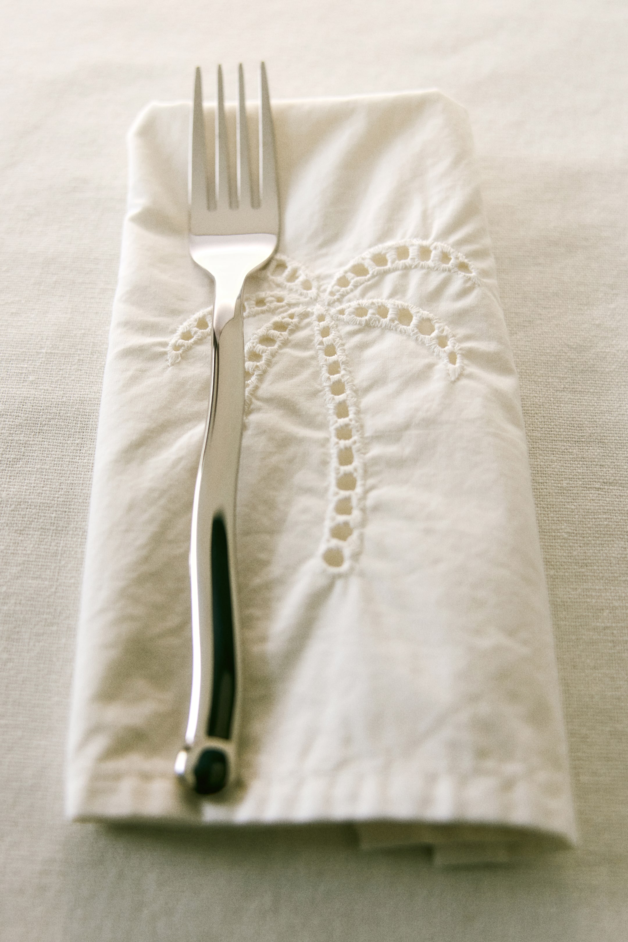 View larger image: A polished metal fork is lying vertically, showcasing four tines and a curved handle with a dark accent at its base. It is resting on a folded, off-white fabric serviette featuring an embroidered palm tree design with eyelet details.