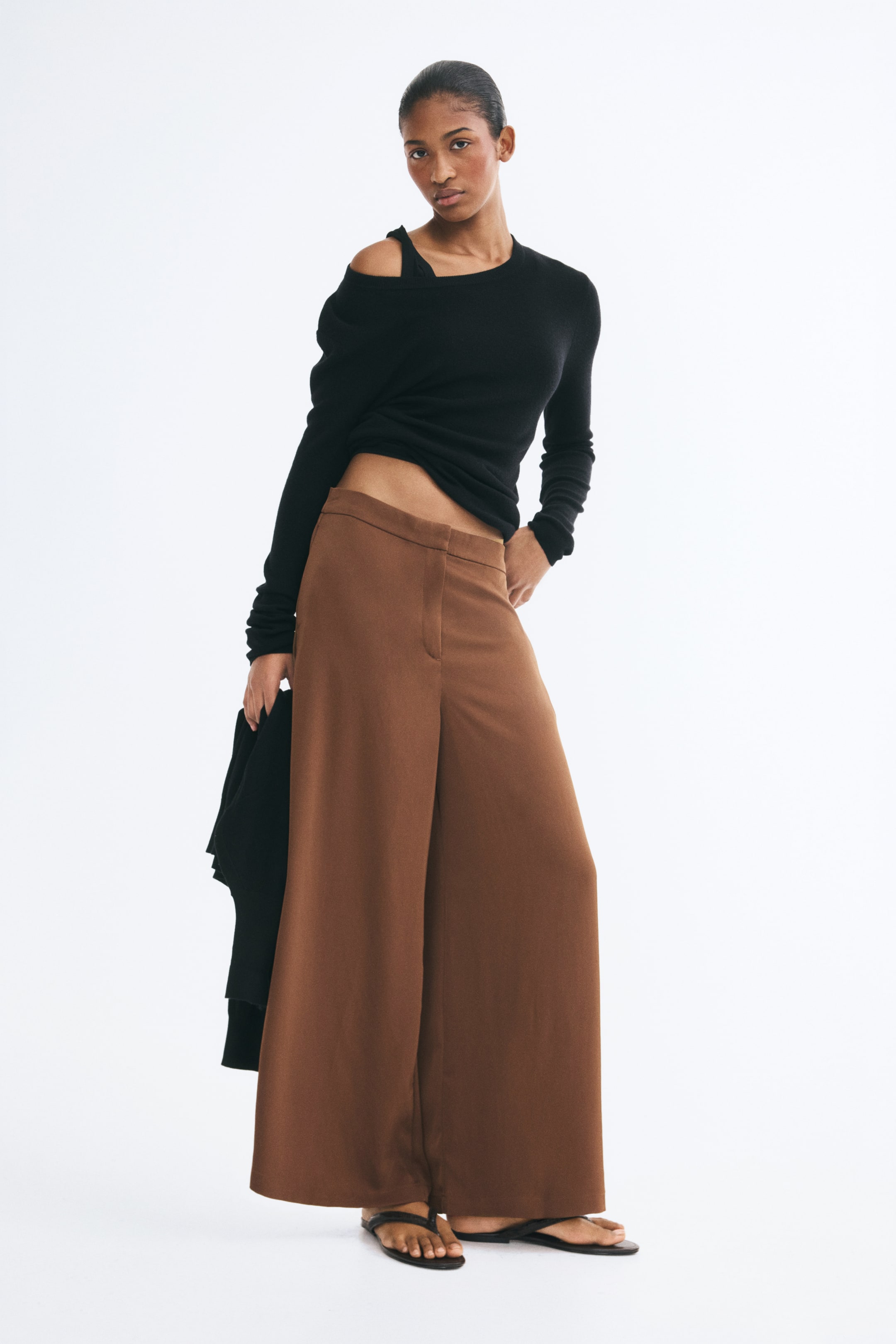 View larger image: Brown wide-leg pants in a woven fabric with a slight sheen are worn by the model, featuring a high waist and a zip fly closure. The pants are styled with a black off-the-shoulder long-sleeve top and flat sandals, while the model is holding a black garment in one hand.