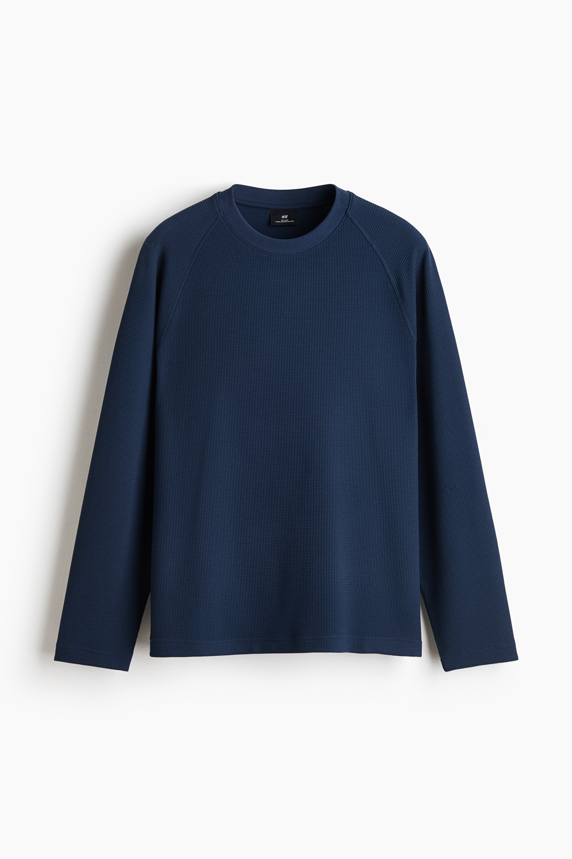 Un t-shirt en jersey gaufrÃ© bleu, Ã  coupe dÃ©contractÃ©e, avec un col rond et des manches raglan longues, portÃ© par le mannequin. Le t-shirt prÃ©sente un ourlet droit et est agencÃ© avec un pantalon gris foncÃ©.