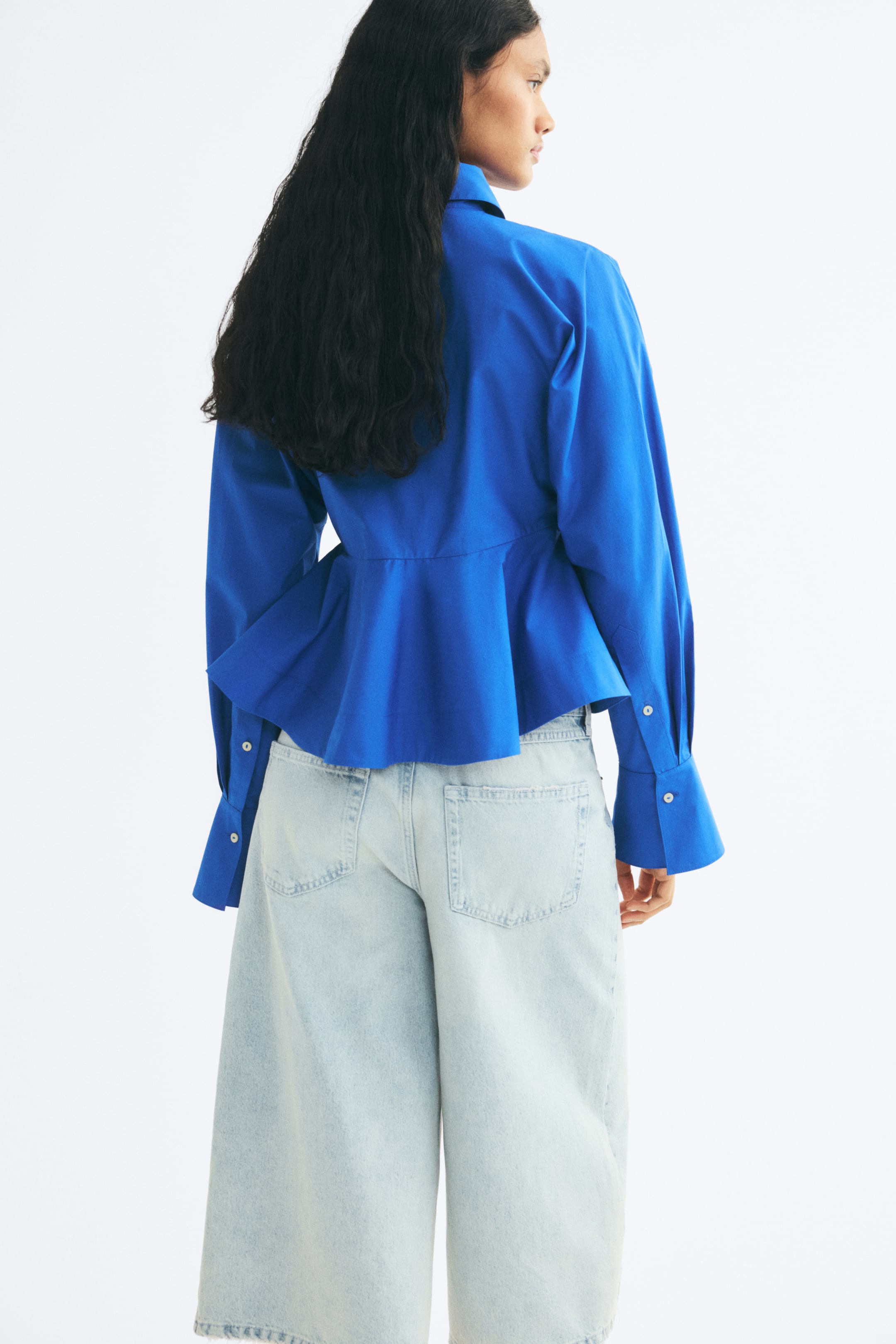View larger image: The model is wearing a bright blue cotton poplin shirt featuring a flared peplum hem and long sleeves with wide, buttoned cuffs. Styled with light-wash, wide-leg denim jeans, the look highlights the shirt’s structured design from the back.