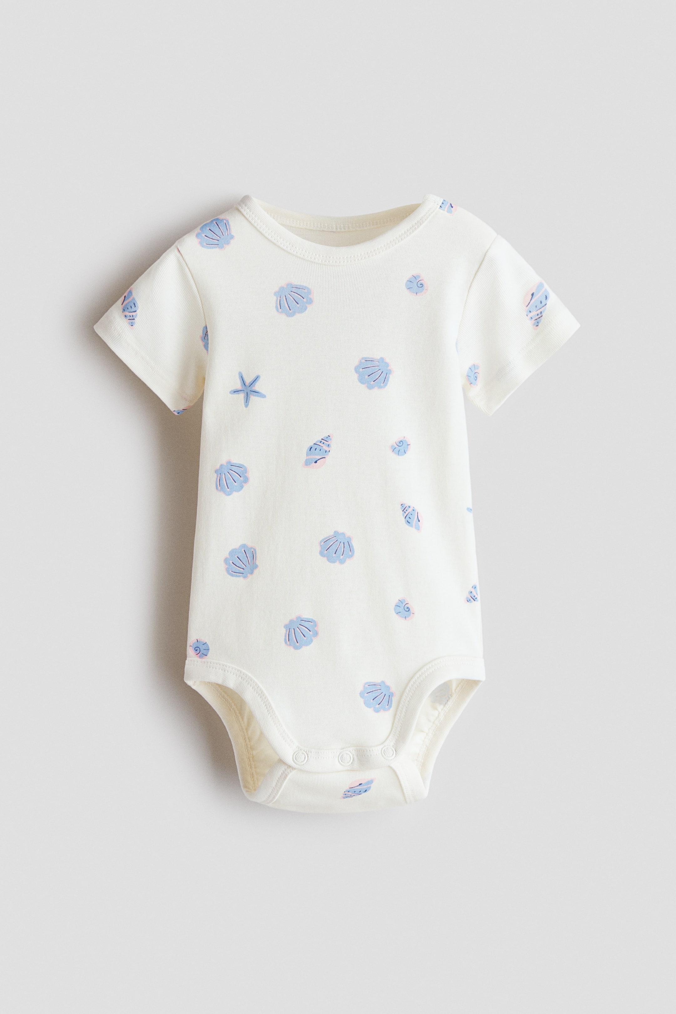 View larger image: A short-sleeved bodysuit in soft cotton jersey is facing front, showcasing an all-over print of light blue seashells and starfish on an off-white base. It features a round, trimmed neckline and fastens with press-studs on the left shoulder and at the crotch.