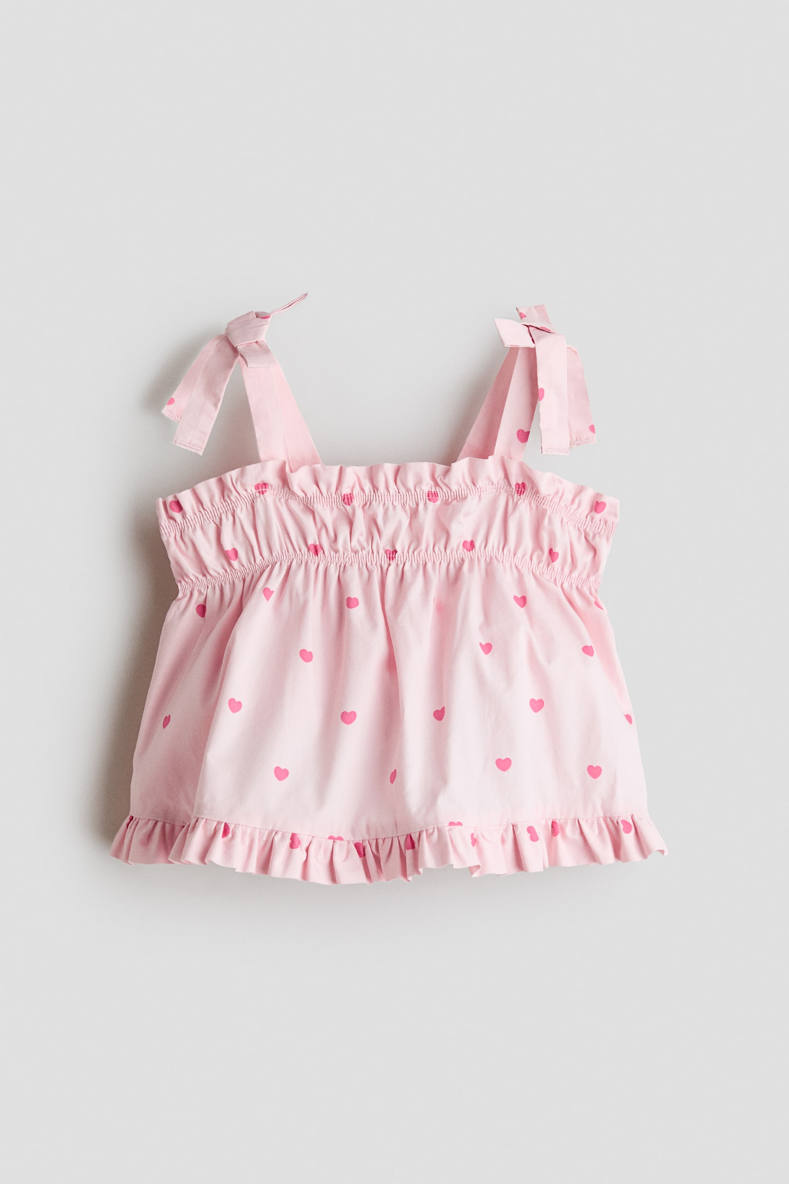 A front-facing light pink sleeveless top features an all-over pink heart print. The top displays tie-shoulder straps, an elasticated smocked chest with a ruffled top edge, and a flared bodice ending in a ruffled hem.