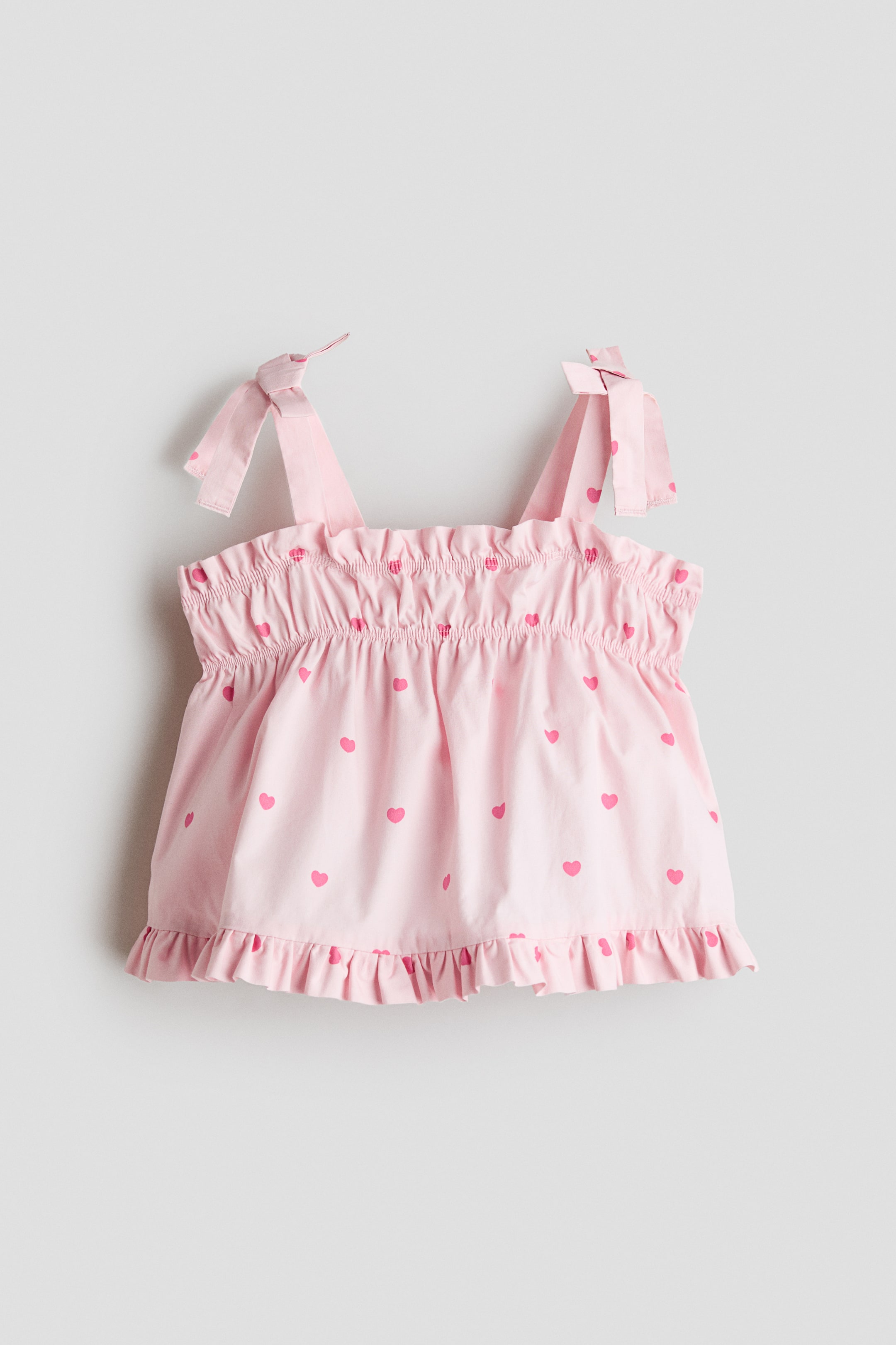 View larger image: A front-facing light pink sleeveless top featuring an all-over pink heart print. The top features tie-shoulder straps, an elasticated smocked chest with a ruffled top edge, and a flared bodice ending in a ruffled hem.