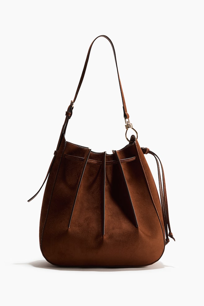 A brown suede bucket bag is facing front, featuring a gathered top with vertical pleats and visible contrast stitching. It is designed with a drawstring closure and a single adjustable shoulder strap. - 1