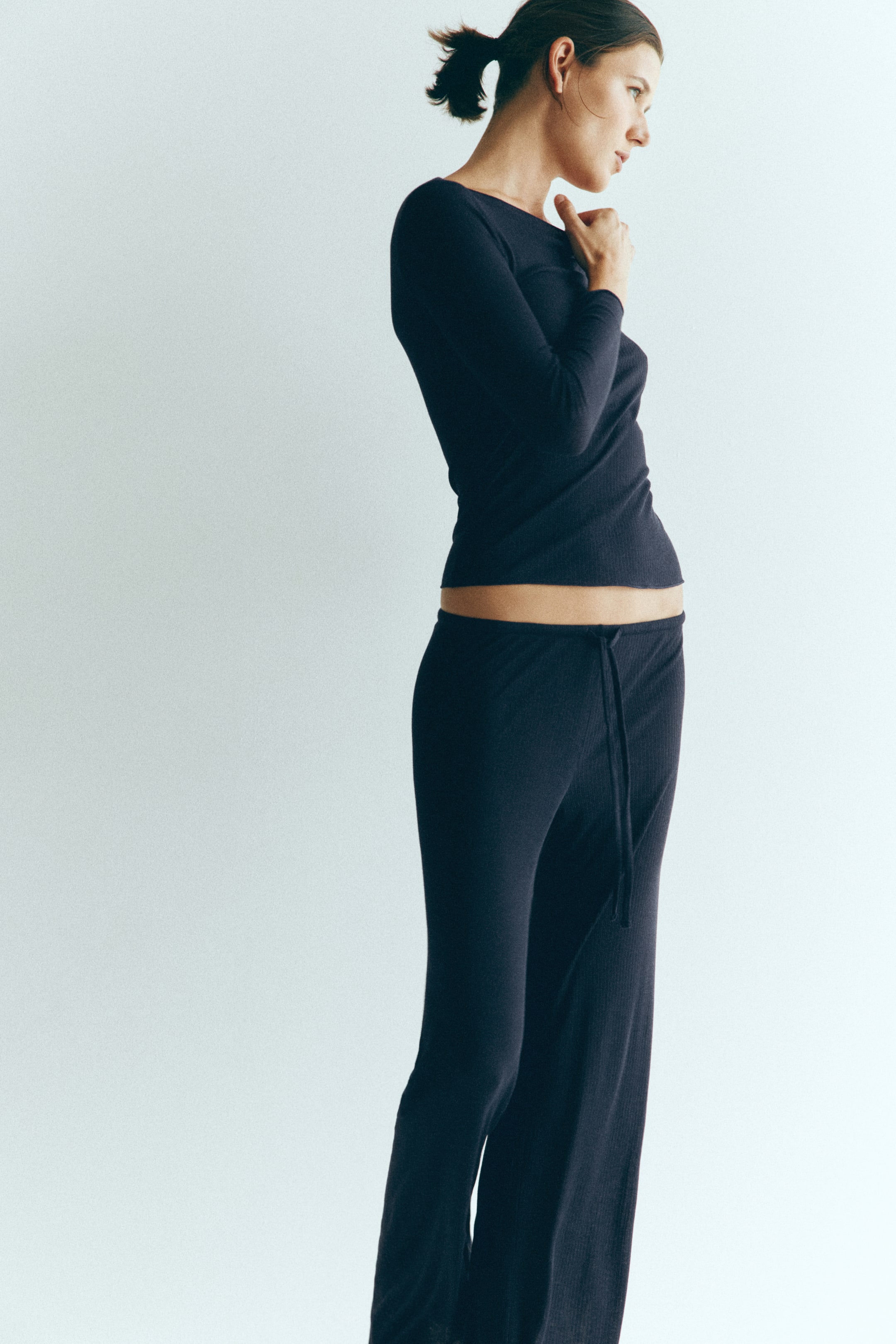 Grösseres Bild ansehen: Ein dunkelblaues, geripptes Jersey-Pyjama-Set wird vom Model getragen, das im Profil steht. Das Set besteht aus einem figurbetonten, langärmeligen Oberteil mit U-Boot-Ausschnitt und einer dazu passenden weit geschnittenen Hose mit Kordelzugbund.