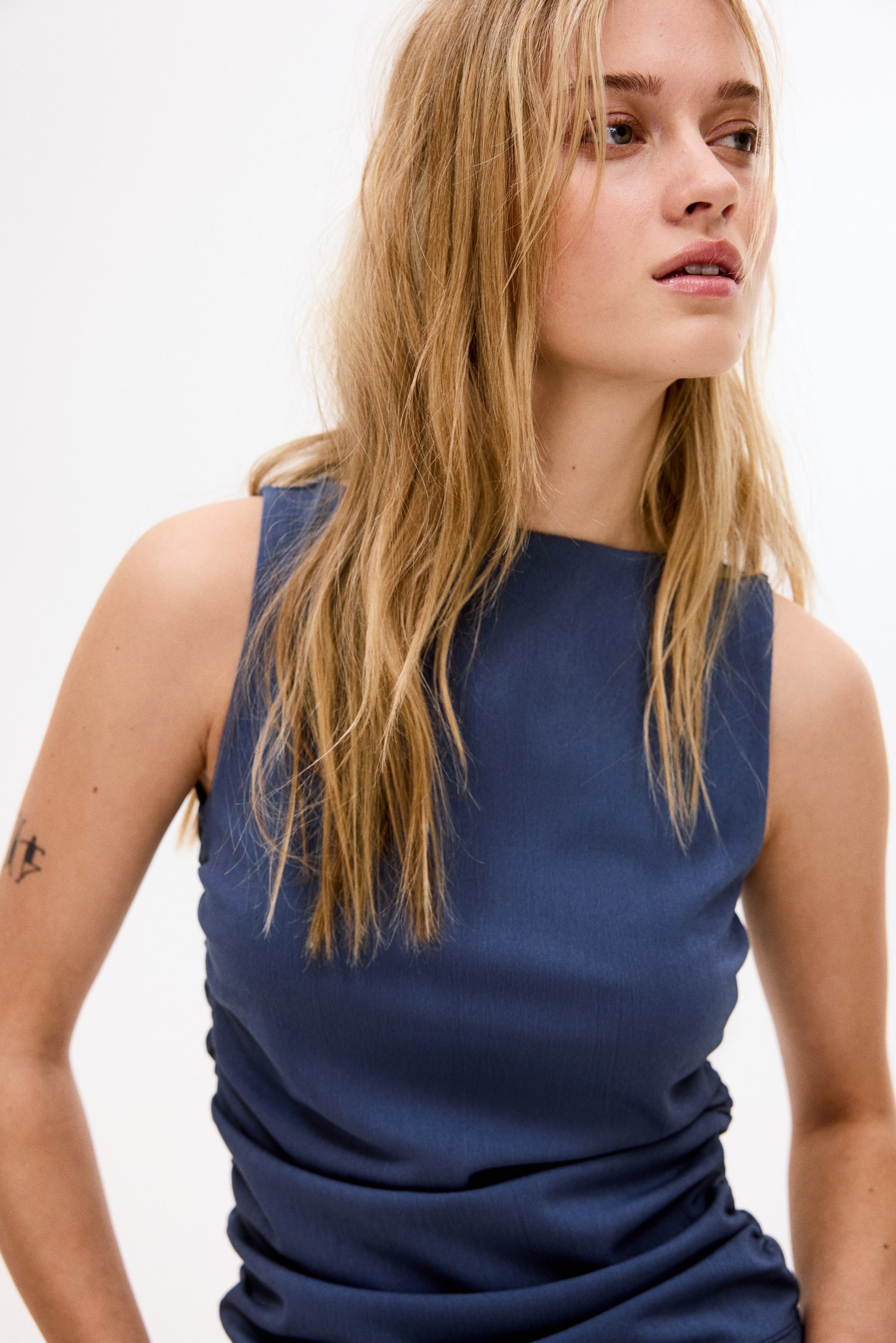 Visa större bild: Den mörkblå ärmlösa toppen har en hög krage och en åtsittande silhuett, med rynkningar på höger sida. Den presenteras från en framsidesvinkel, vilket framhäver dess texturerade tyg.