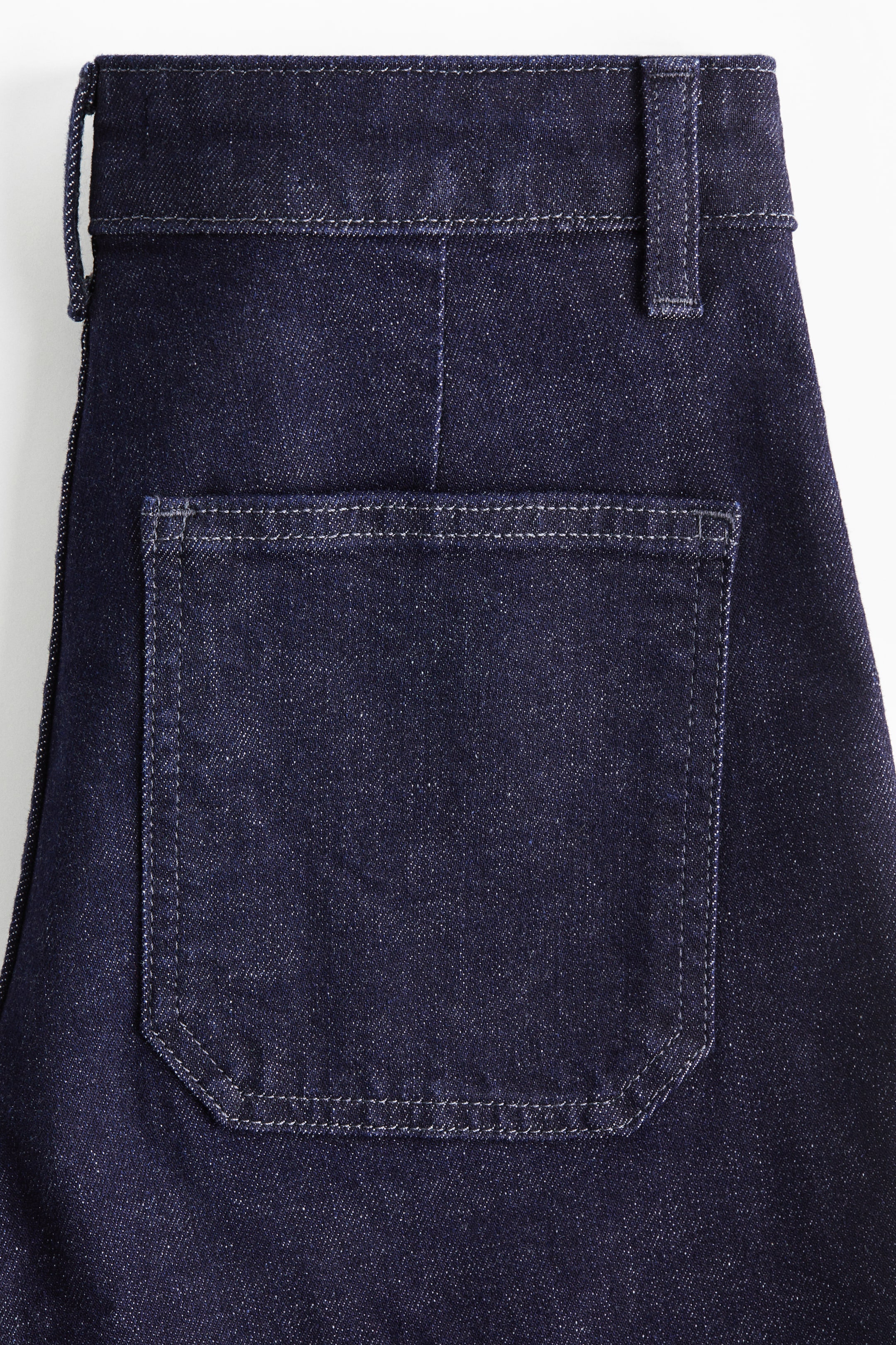 Se større billede: Mørkeblå denimjeans har en rektangulær baglomme med kontrasterende lysfarvet syning, inklusive 'V'-formede forstærkninger i de nederste hjørner. Linningen har bæltestropper og en central bagstøm.