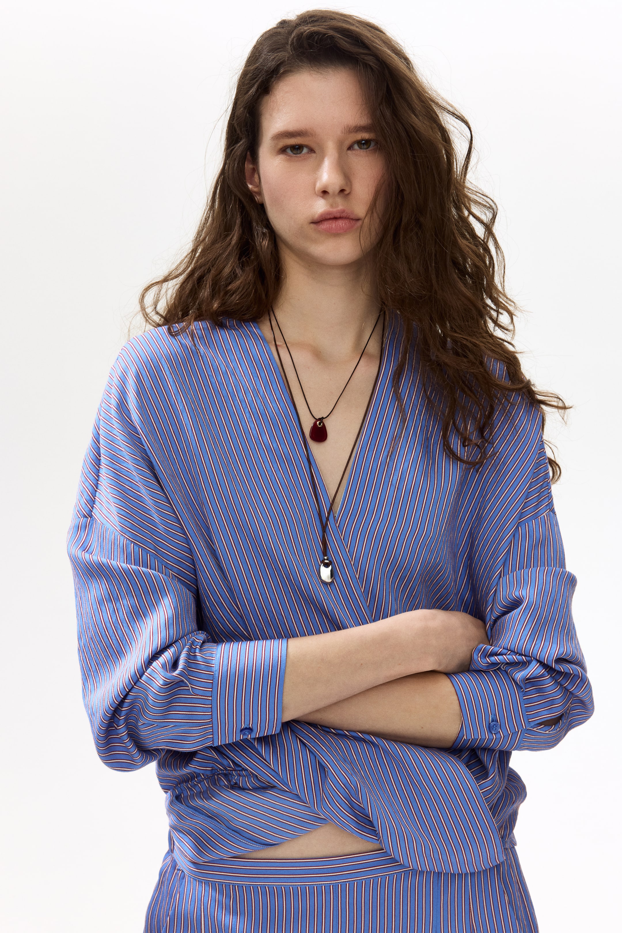 View larger image: A blue wrap shirt adorned with delicate white and red vertical stripes is worn, showcasing a V-neckline and long sleeves with buttoned cuffs. The shirt cinches at the waist, revealing the midriff, and is paired with a coordinating striped bottom.