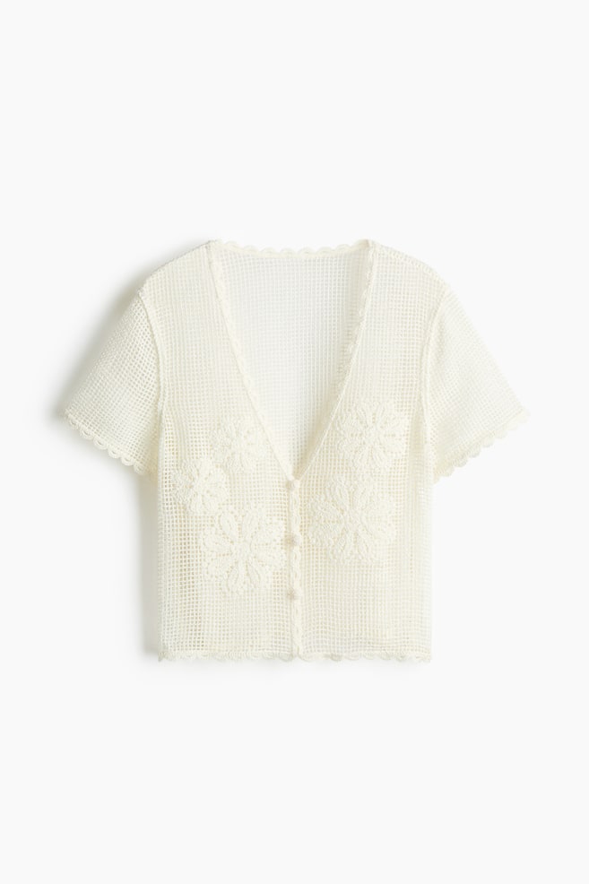 A short, fitted, cream-coloured beach top is presented FRONT, featuring a crochet-look knit with floral motifs on the front. It is designed with a deep V-neckline, covered buttons down the front, short sleeves, and scalloped edges. - 1