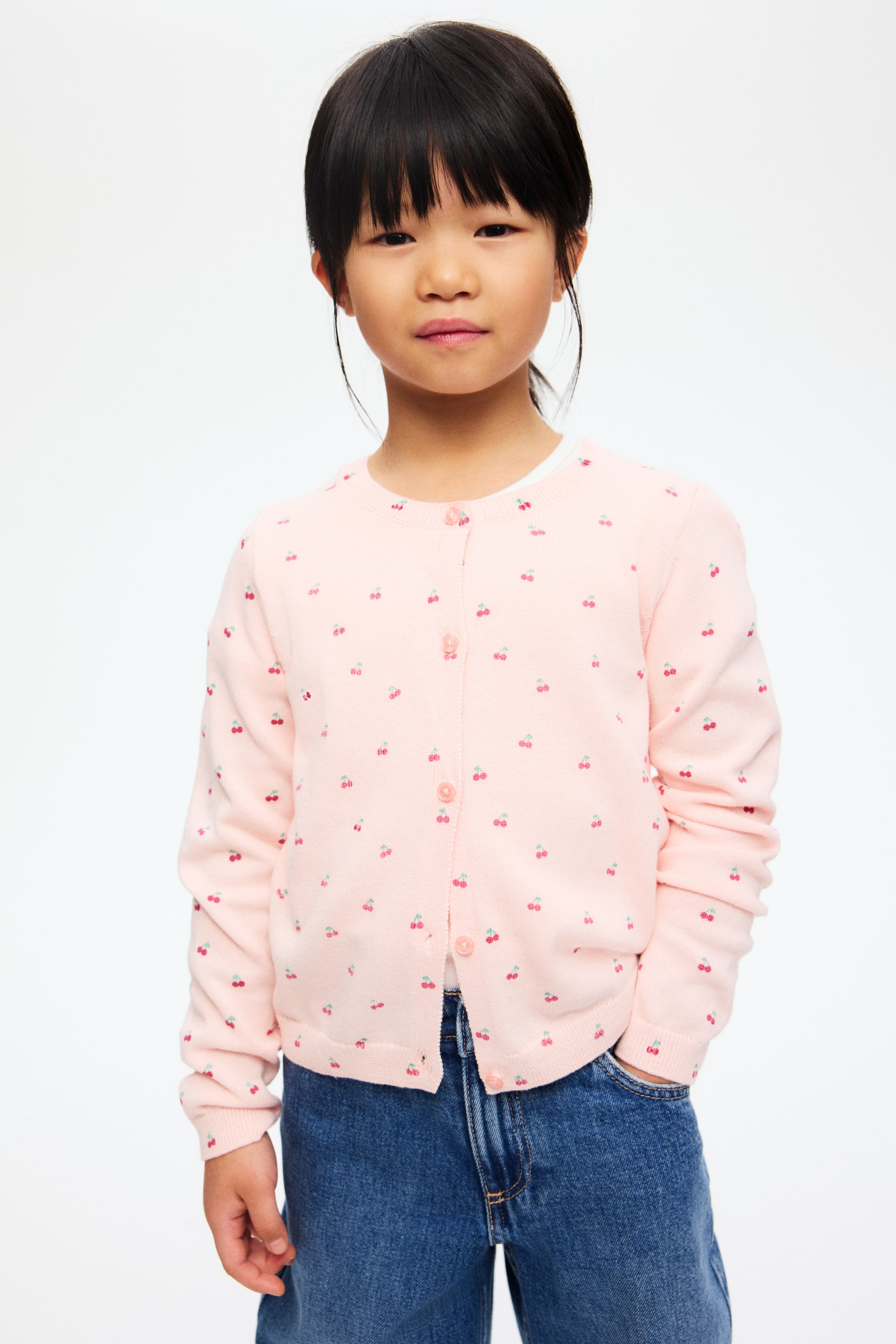 Agrandir l'image: Le cardigan rose clair, orné d'un motif de cerises dispersées, est superposé sur un haut blanc et est présenté de face. Il est conçu avec un col rond, une fermeture à boutons sur le devant avec des boutons roses assortis, et est fini avec des poignets et un ourlet côtelés.