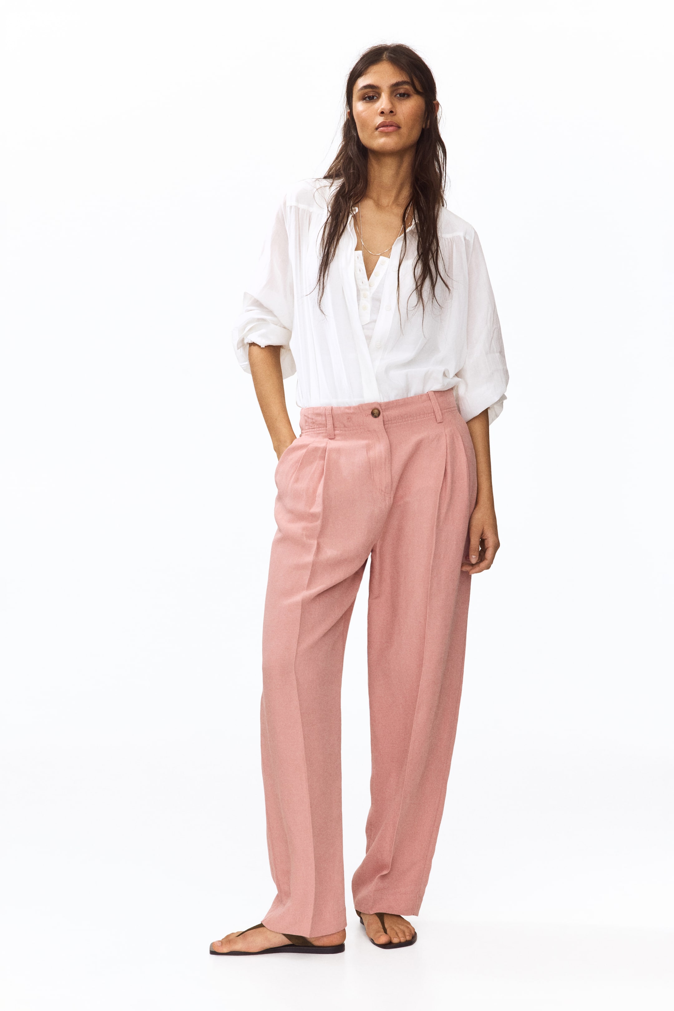 View larger image: "Pink wide-leg trousers feature a high-waisted design with a single button closure and double front pleats. Crafted from textured fabric, they offer a relaxed fit and come with side pockets, styled with a white shirt tucked in."