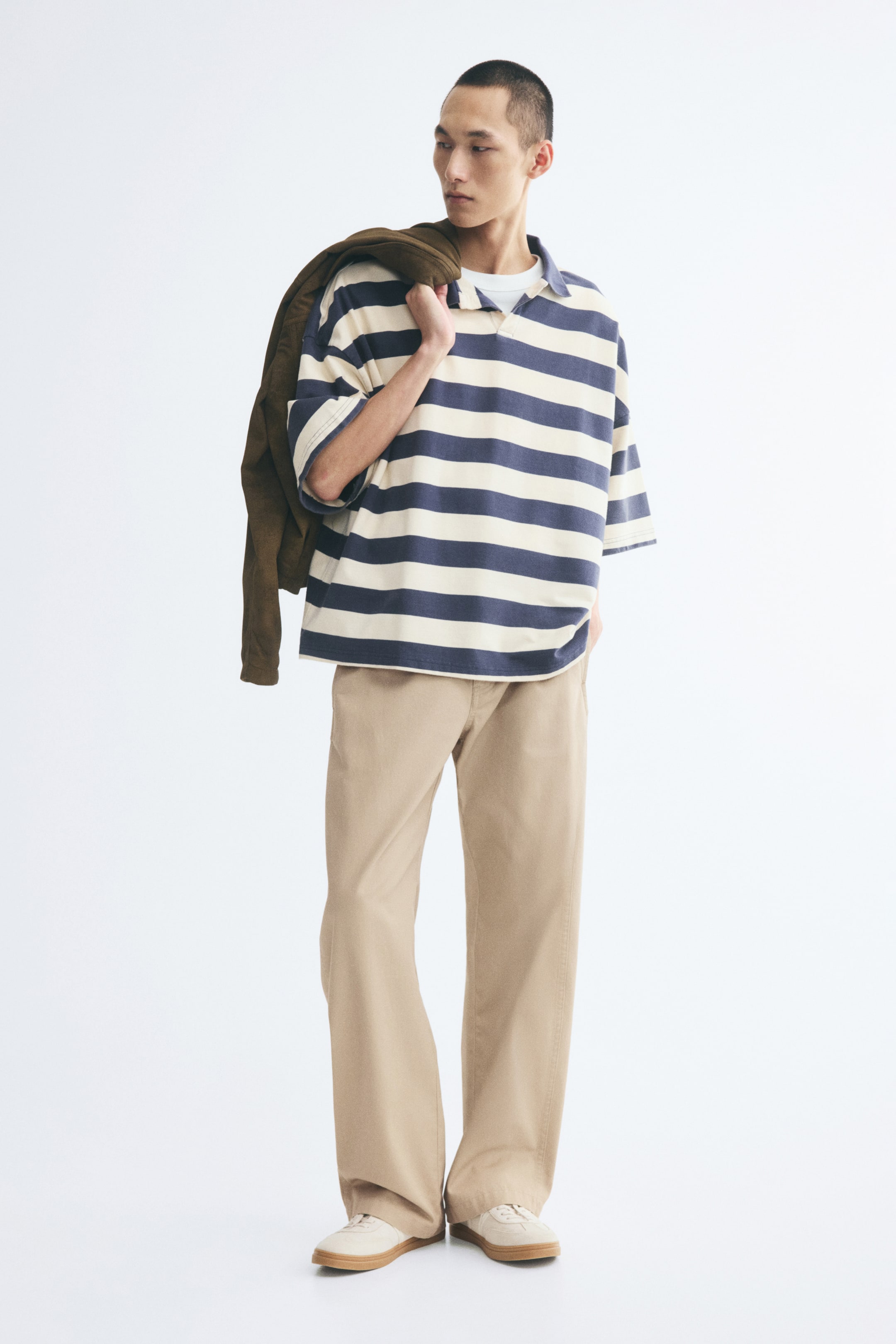 View larger image: Beige cotton twill chinos featuring a relaxed fit and barrel-shaped legs are being worn by the model. The model is styling the trousers with a blue and cream striped polo shirt and light-coloured sneakers while carrying a brown jacket over one shoulder.