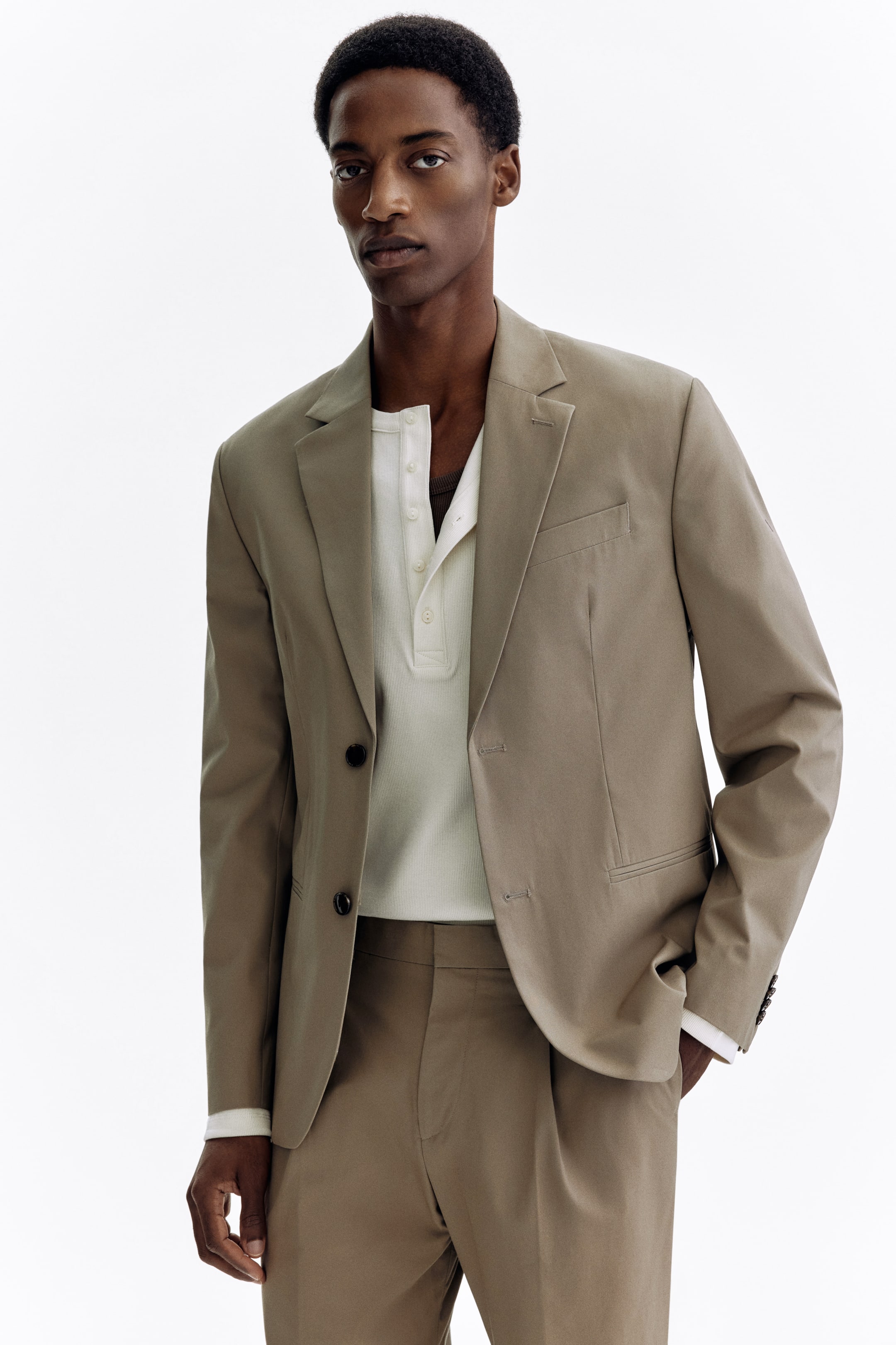 View larger image: A taupe suit, comprising a single-breasted blazer and matching pleated trousers, is showcased from a slightly angled front perspective. The blazer features notch lapels, a two-button closure with one button fastened, a welt chest pocket, and flap pockets, while each cuff is detailed with four buttons. A white Henley shirt is layered underneath, visible at the blazer's open front.