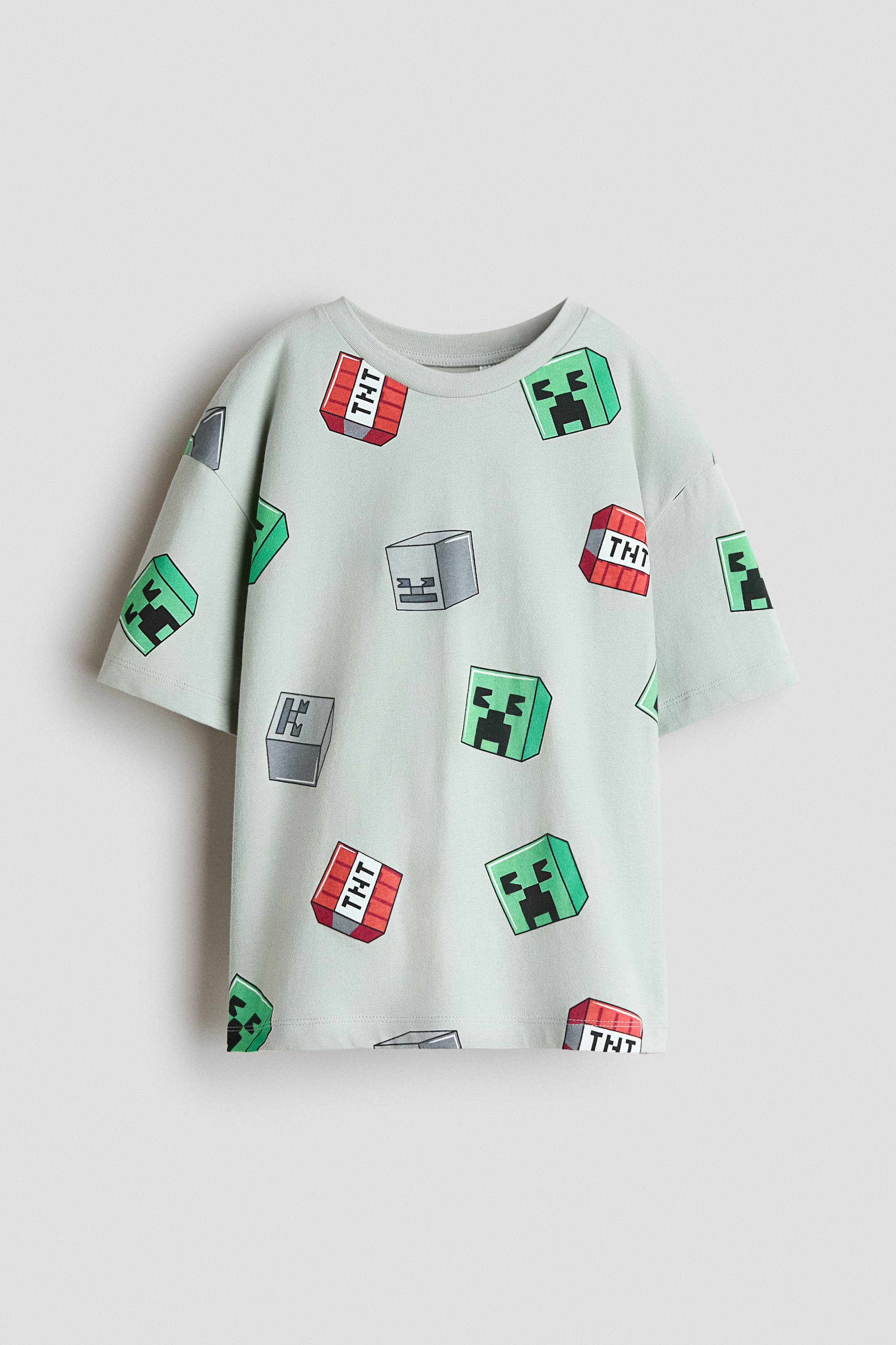 Se større billede: En lysegrøn T-shirt med korte ærmer har rund hals og et heldækkende print af grønne Creeper-ansigter, røde TNT-blokke og grå blokke. T-shirten ligger fladt og er vist forfra.