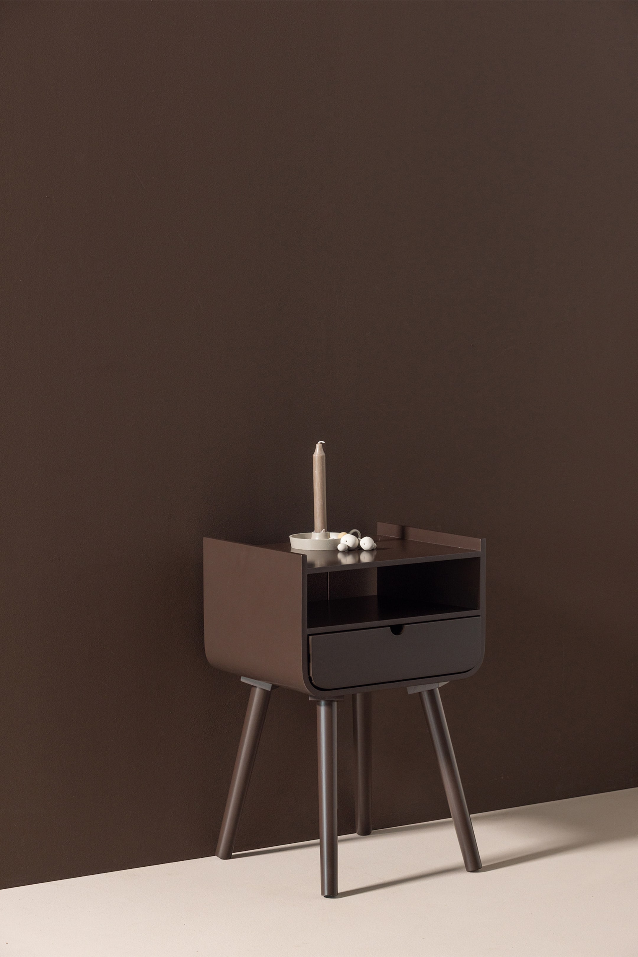 Agrandir l'image: Une table de chevet marron foncé présente son côté avant et gauche, avec une étagère ouverte et un tiroir marron plus foncé aux bords inférieurs arrondis. Elle repose sur quatre pieds effilés et inclinés, tandis qu'une bougie et de petits objets décoratifs blancs sont posés sur sa surface supérieure.
