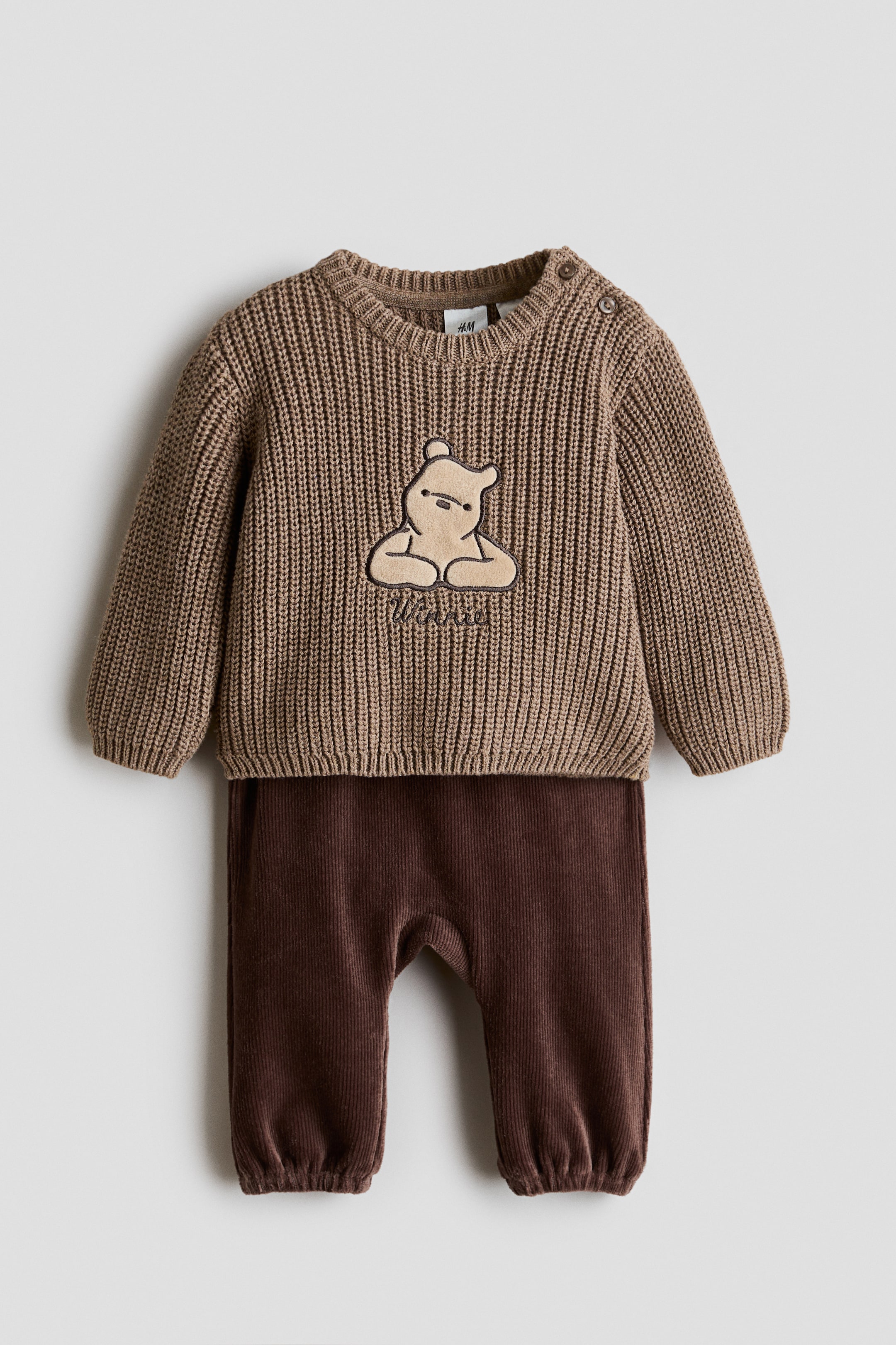 Größeres Bild anzeigen: Ein braunes geripptes Strickpullover- und dunkelbraunes Cordhosen-Set wird präsentiert. Der Pullover zeigt einen beige bestickten Winnie-the-Pooh-Aufnäher auf der Vorderseite mit dem Text „Winnie“ darunter und zwei Knöpfen auf der linken Schulter, während die Cordhosen geraffte Knöchel aufweisen.