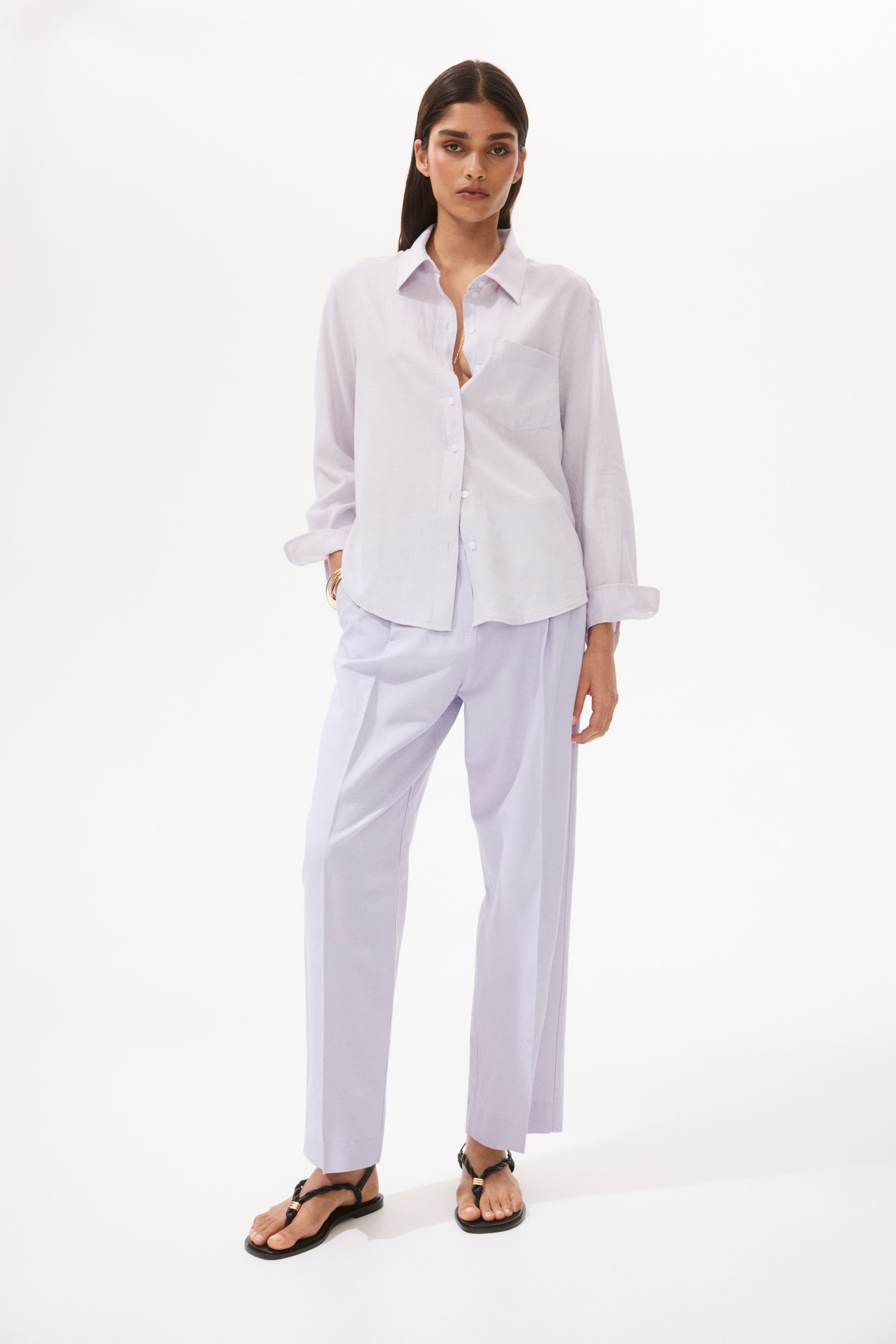 View larger image: A light lavender button-up shirt, featuring a pointed collar, chest patch pocket, and rolled-up long sleeves, is styled loosely tucked into matching light lavender wide-leg trousers with front pleats. Black braided flat sandals adorned with gold-toned embellishments complete the look.
