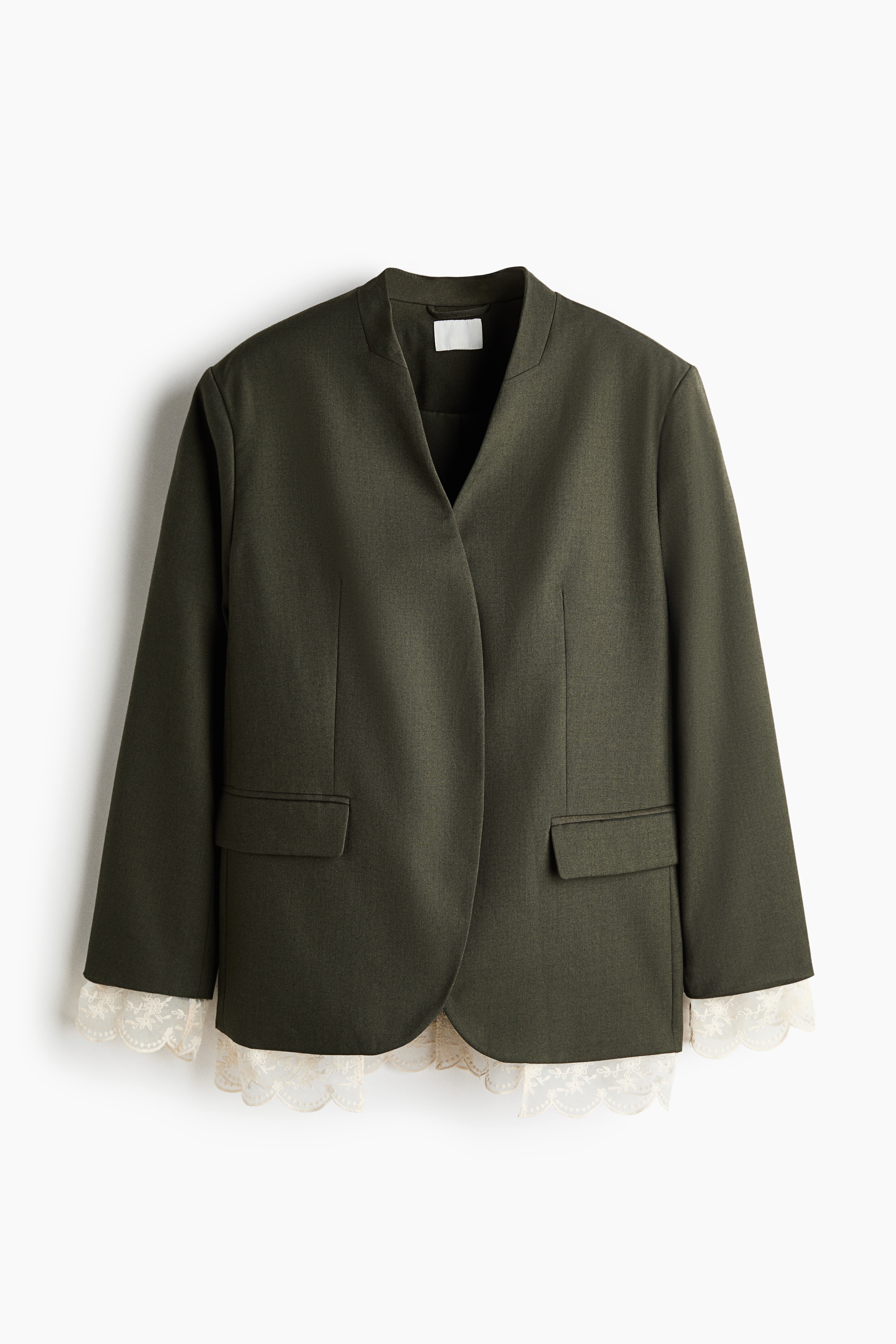 A dark olive green, lapelless blazer is worn by the model over a draped olive-coloured top and matching pleated skirt. The open-front blazer features structured shoulders, a gold brooch pinned to the chest, and delicate white scalloped lace trim extending from the cuffs.