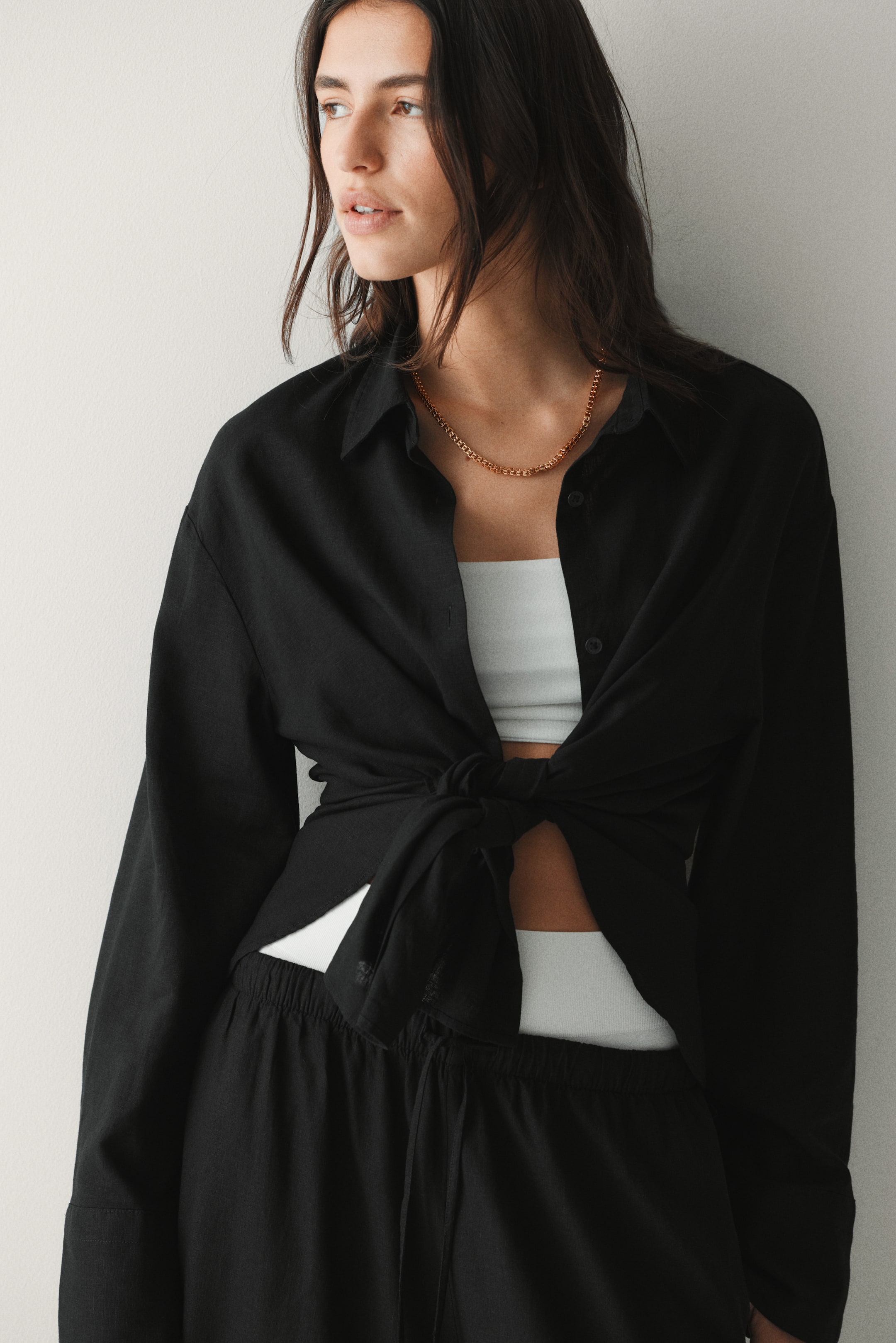 View larger image: A black long-sleeved shirt with a pointed collar is worn open and tied at the waist, layered over a white bandeau top, and styled with black drawstring trousers. A gold chain necklace is also visible around the neck.