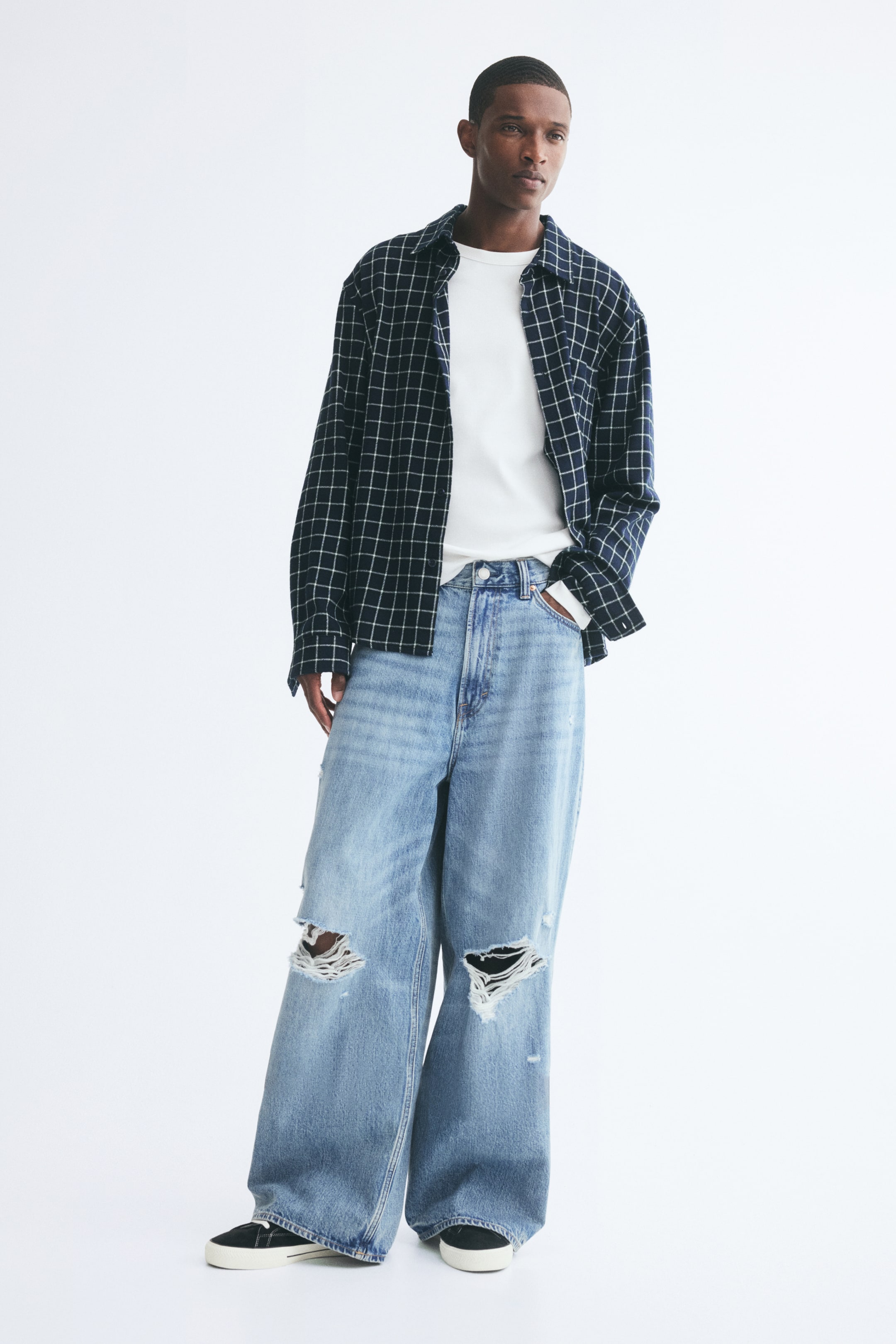 View larger image: Light blue wide-leg denim jeans featuring distressed knee rips and a baggy fit are worn by the model, who is standing with one hand tucked into the pocket. The jeans are styled with a tucked-in white T-shirt, an open navy-and-white grid-patterned long-sleeve shirt, and black sneakers with white soles.