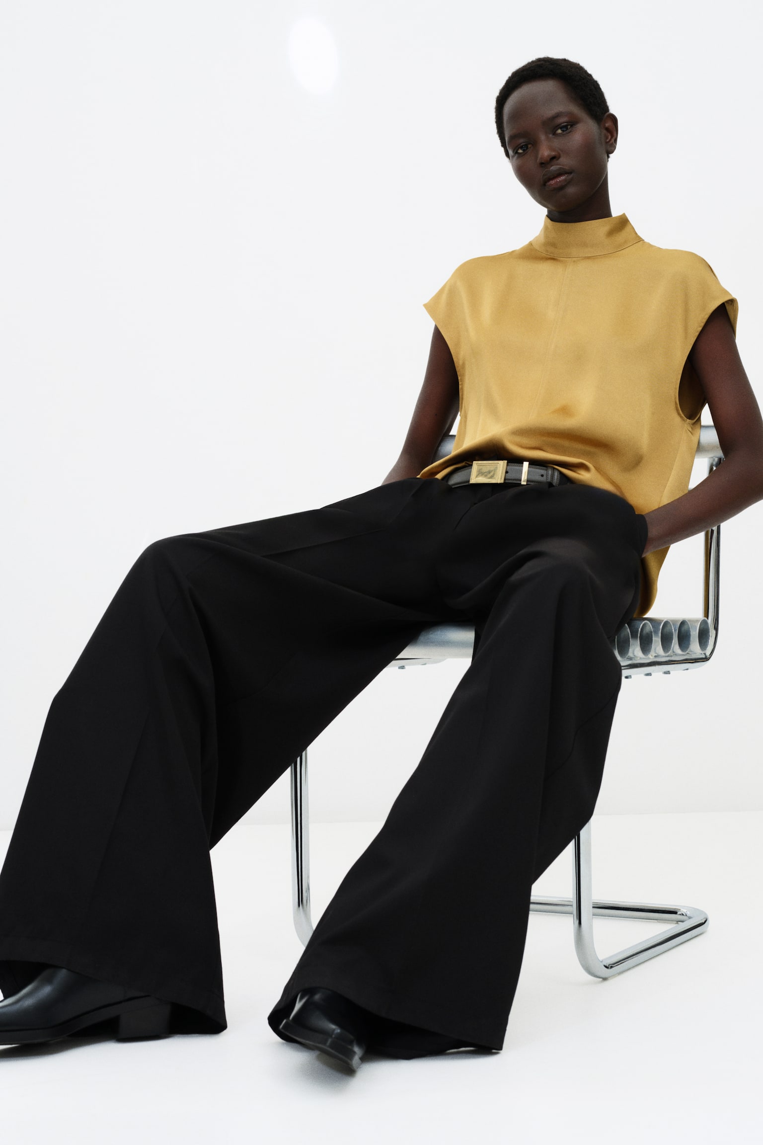 A gold-coloured satin blouse featuring a high stand collar, cap sleeves, and a visible centre seam is being worn by the model. The blouse is tucked into black wide-leg trousers styled with a black belt. - 1