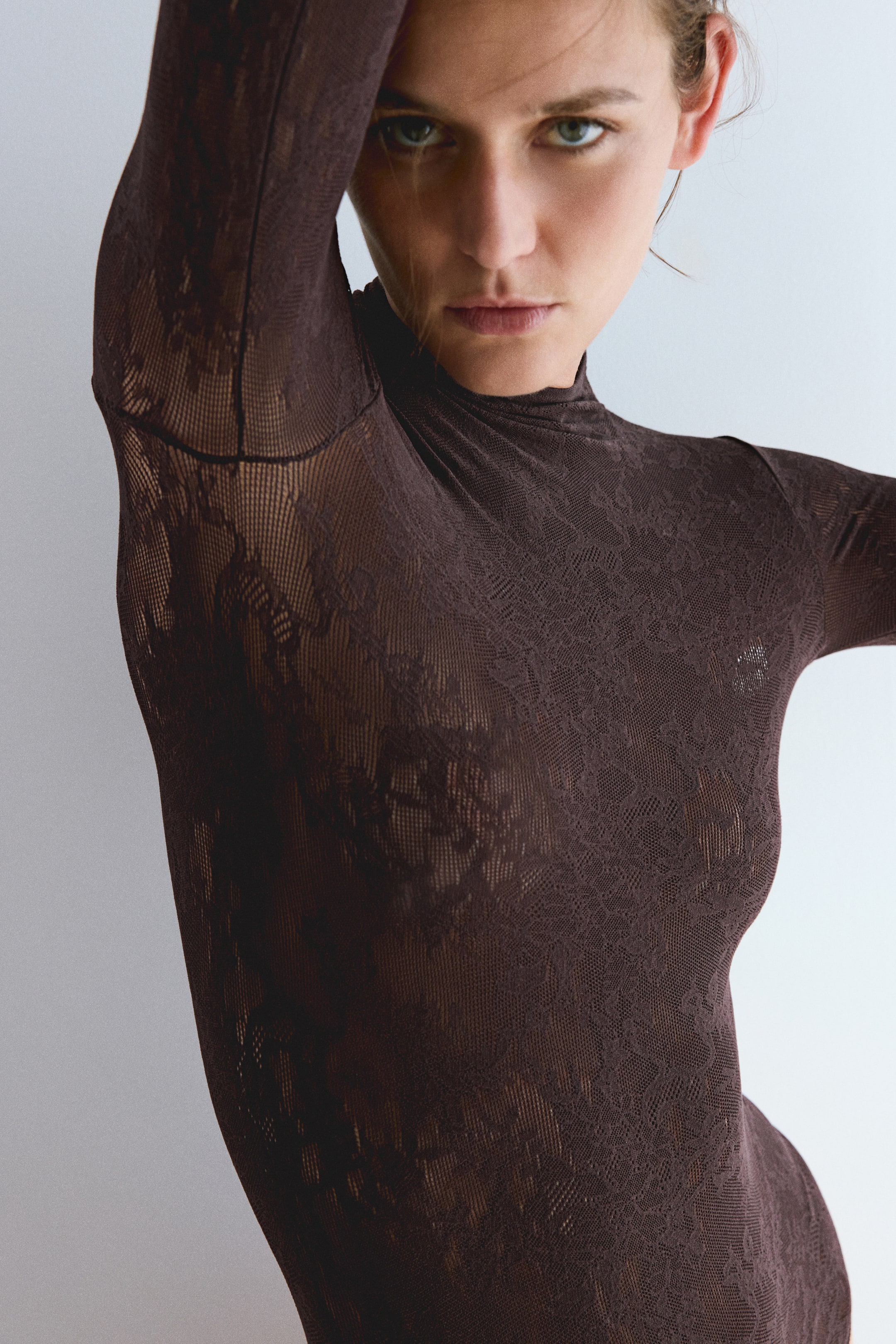 View larger image: A dark brown lace bodysuit with a high neck and long sleeves is worn by the model. The semi-sheer patterned fabric is shown in a close-up as the model poses with one arm raised.