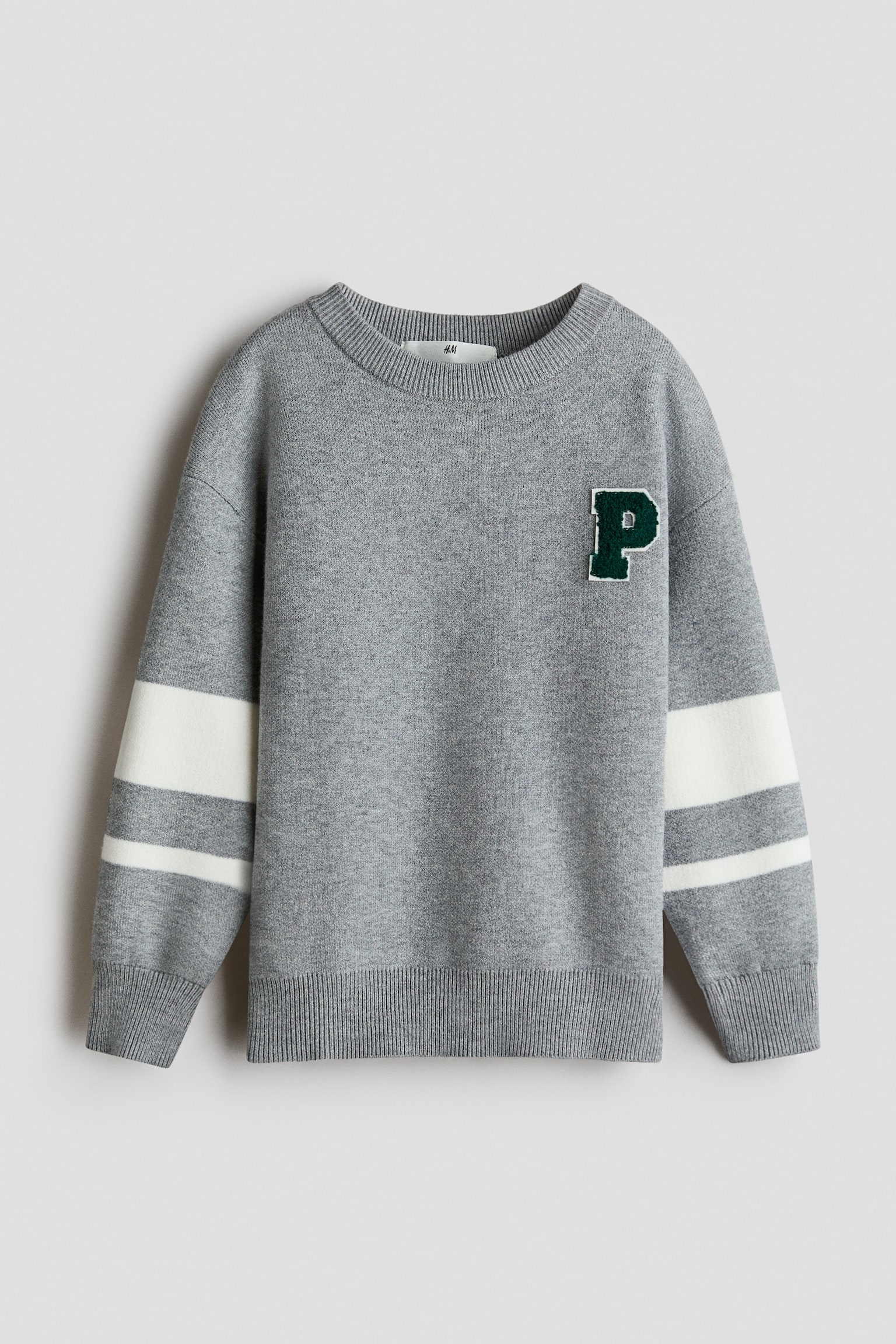 SivÃ½ pletenÃ½ sveter zobrazenÃ½ spredu mÃ¡ na Ä¾avej strane hrude zelenÃº nÃ¡Å¡ivku s pÃ­smenom âPâ v Å¡tÃ½le varsity. Sveter mÃ¡ spustenÃ© ramenÃ¡, dva biele vodorovnÃ© pruhy na kaÅ¾dom rukÃ¡ve a rebrovanÃ© lemy okolo okrÃºhleho vÃ½strihu, manÅ¾iet a spodnÃ©ho okraja.